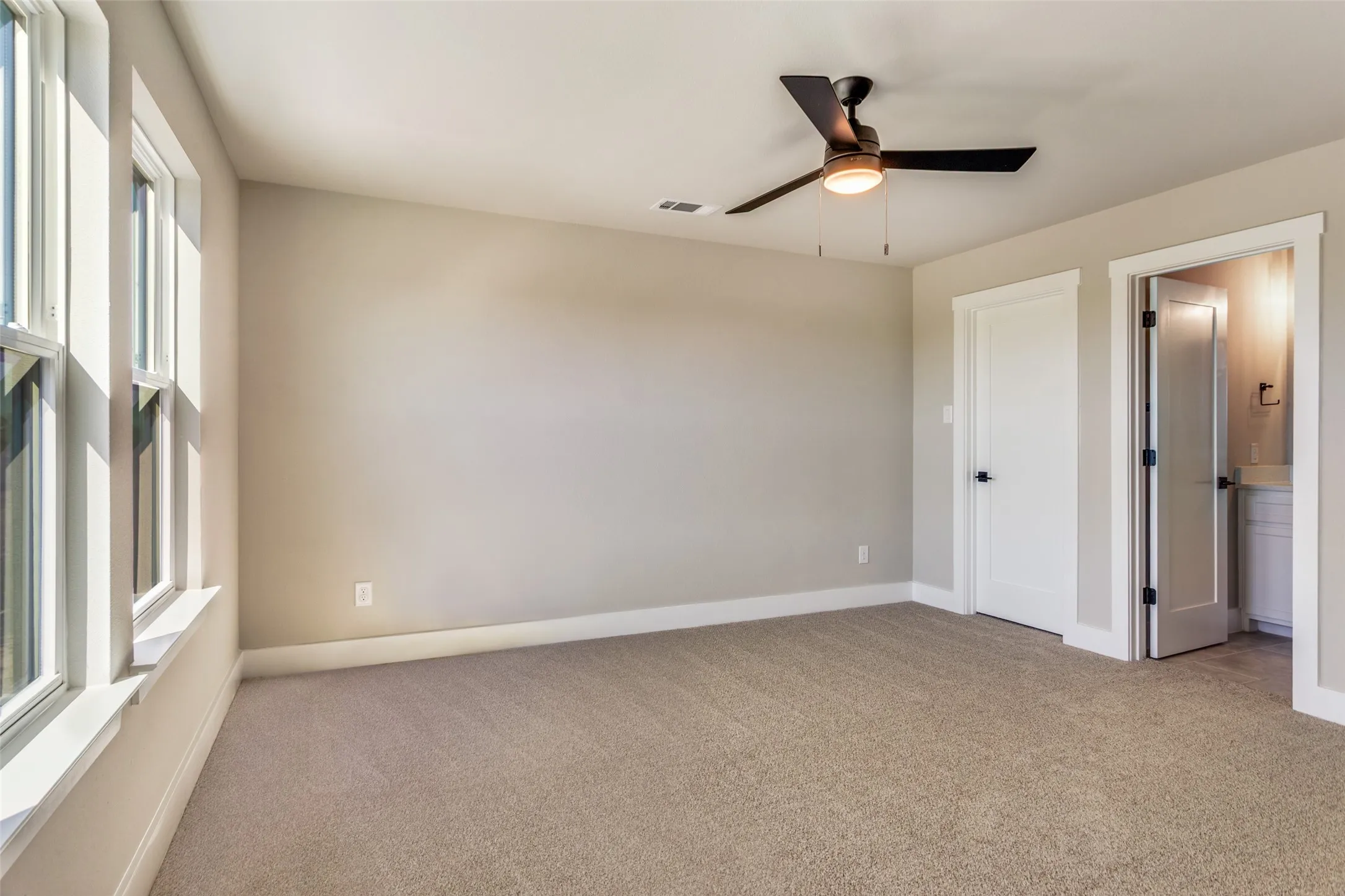 Neutral tone wall paint, carpeted flooring, multiple windows, ceiling fan with integrated lighting, and a white paneled door