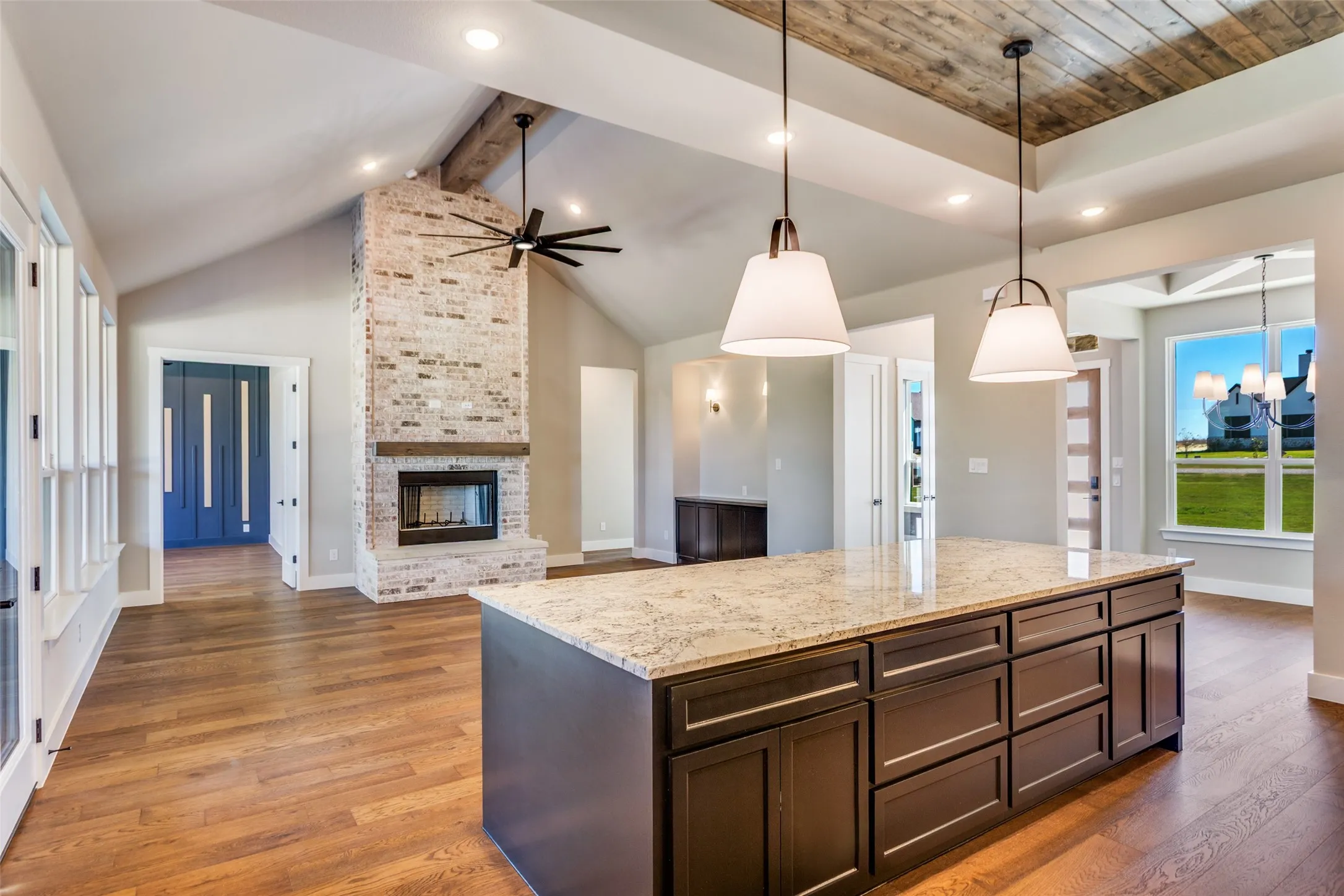 Open-concept living space with vaulted ceilings, a prominent brick fireplace, wood-finish flooring, a kitchen island with stone countertop, and decorative pendant lighting