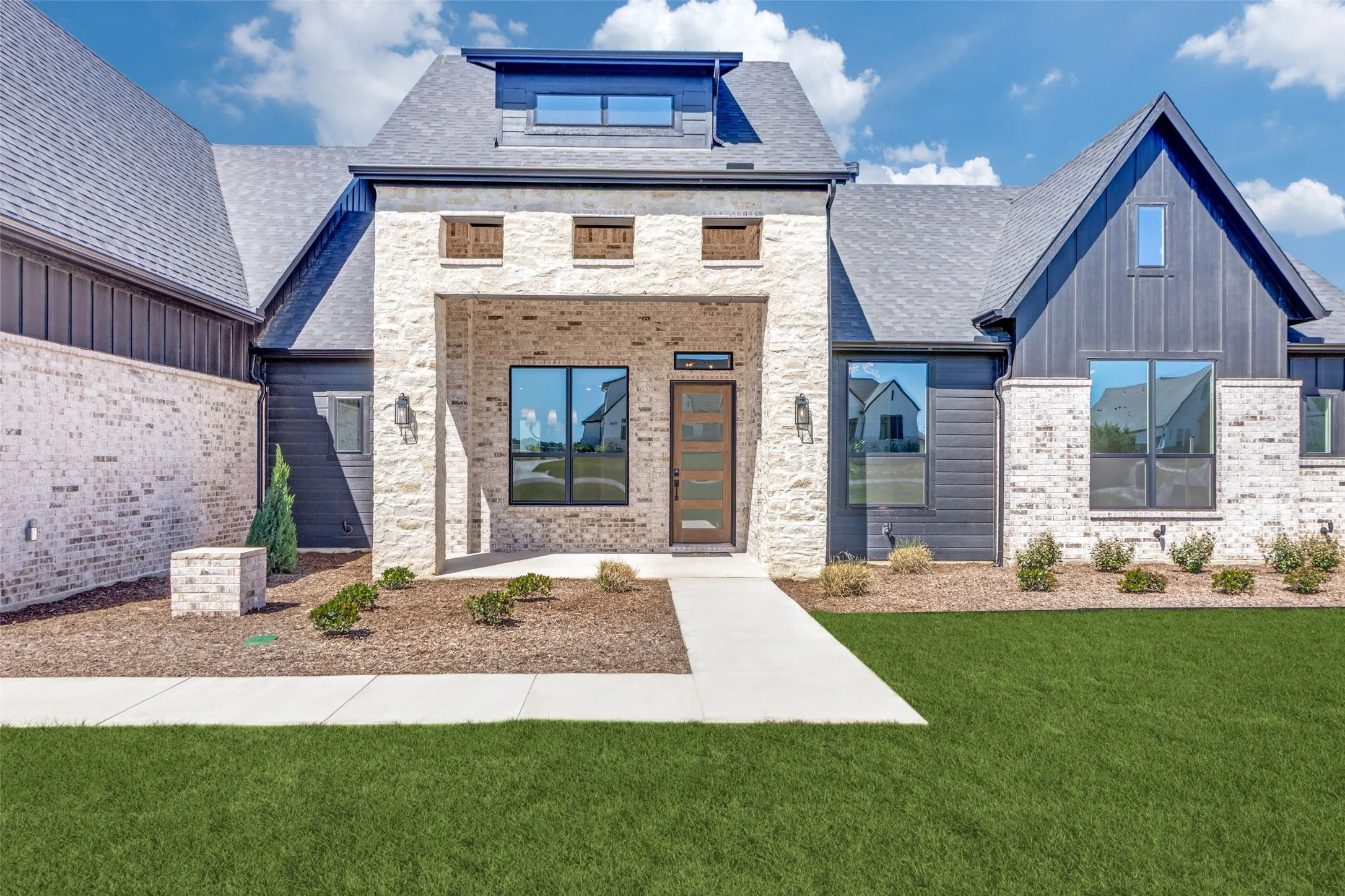 Modern facade featuring light-colored brick and dark vertical siding, an inviting covered entry, a sophisticated front door with multiple glass panes, dormer windows, and a well-maintained lawn