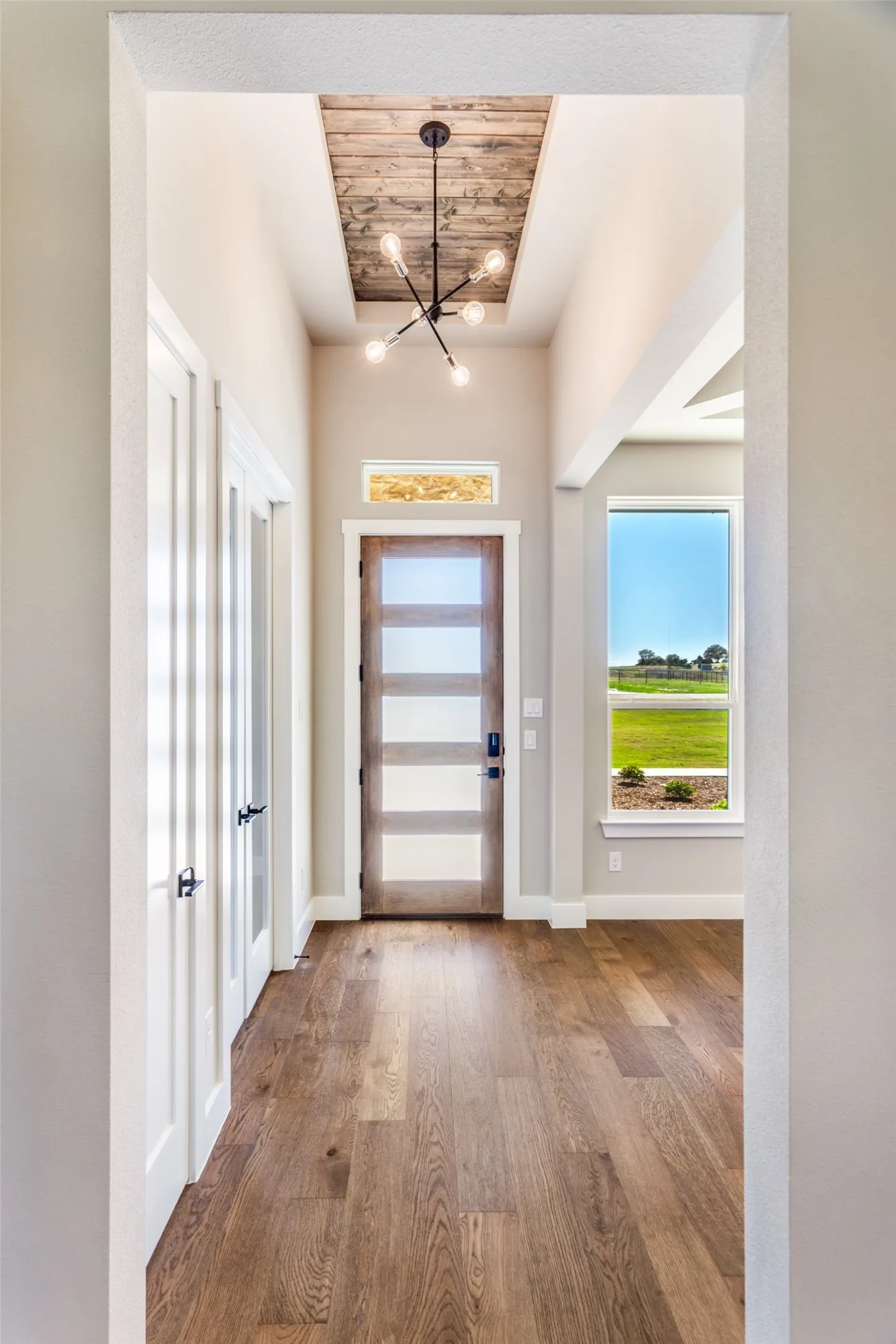 Wood-finish flooring throughout, modern entry door with frosted glass panels, wood plank ceiling accent, and contemporary chandelier