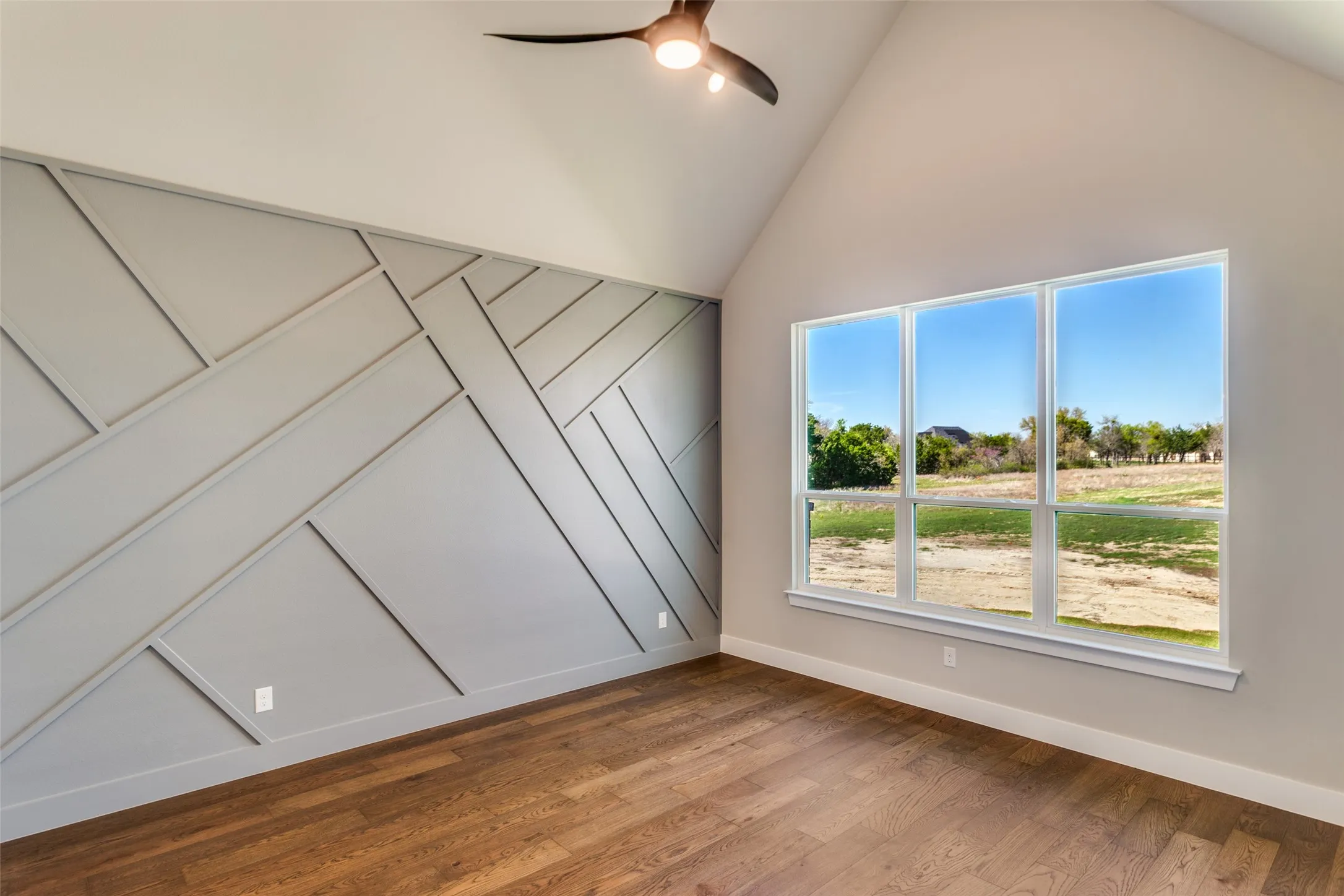 Vaulted ceiling with a contemporary ceiling fan, accent wall with geometric paneling, expansive windows, and wood-finish flooring