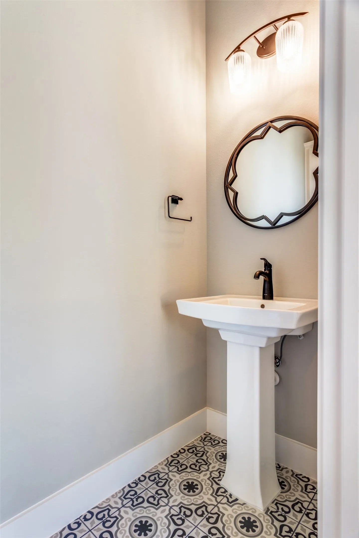 Pedestal sink with black hardware, decorative floor tiling, an ornate mirror, and a two-light fixture