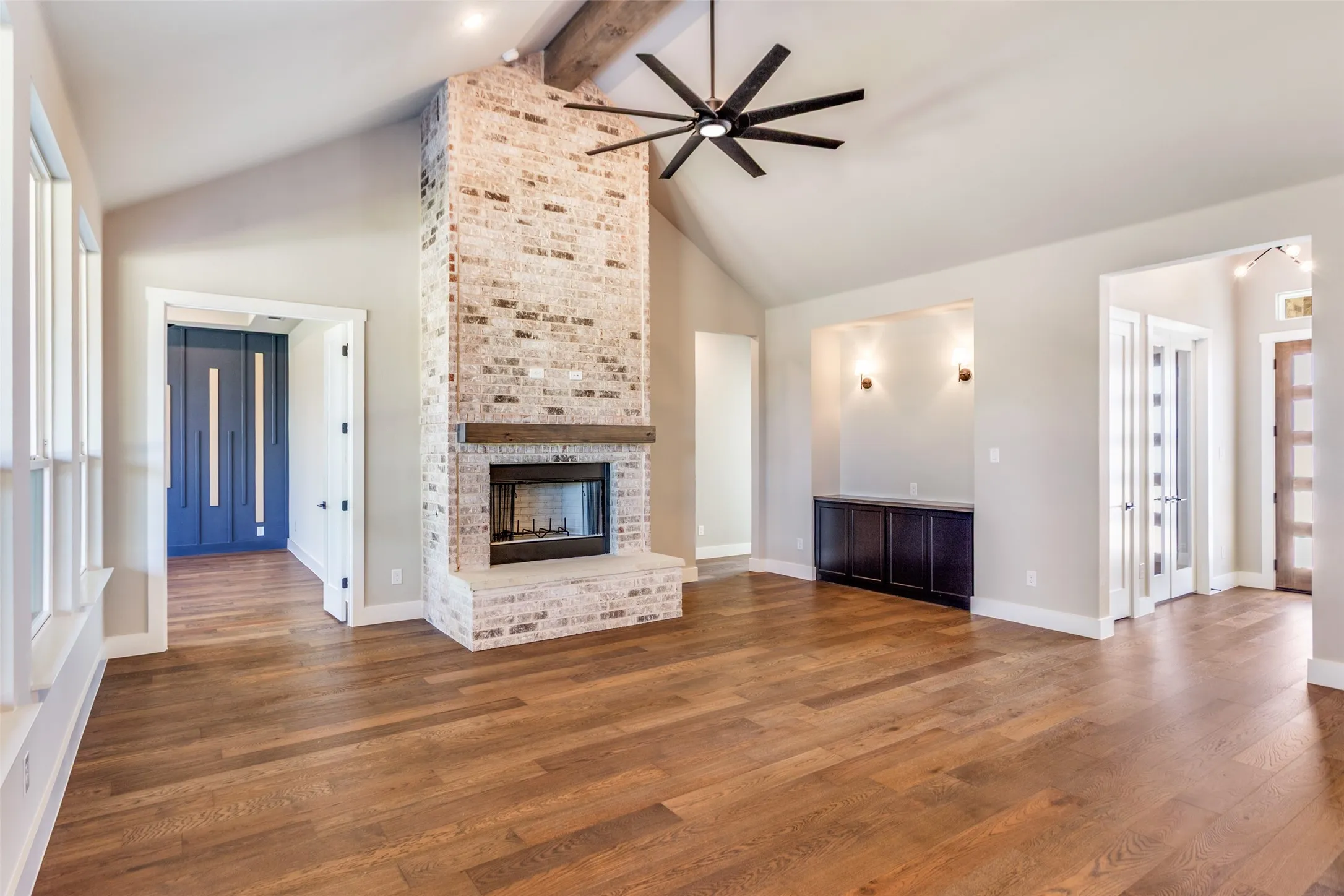 Vaulted ceiling with exposed wood beam, wood-finish flooring, brick fireplace with wood mantel, built-in cabinetry, and modern ceiling fan