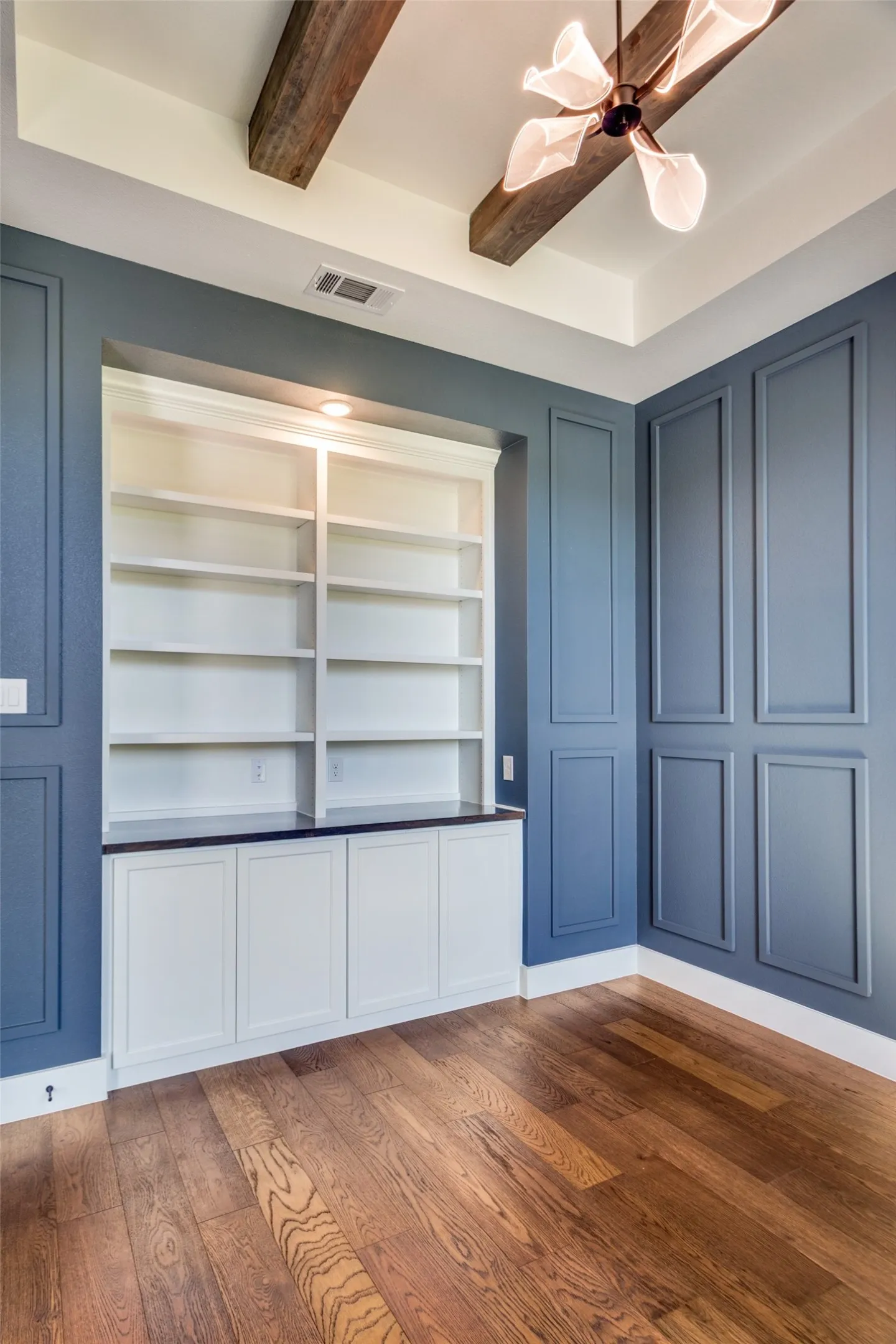 Wood-finish flooring with built-in shelving, paneled walls, and a tray ceiling with exposed beams
