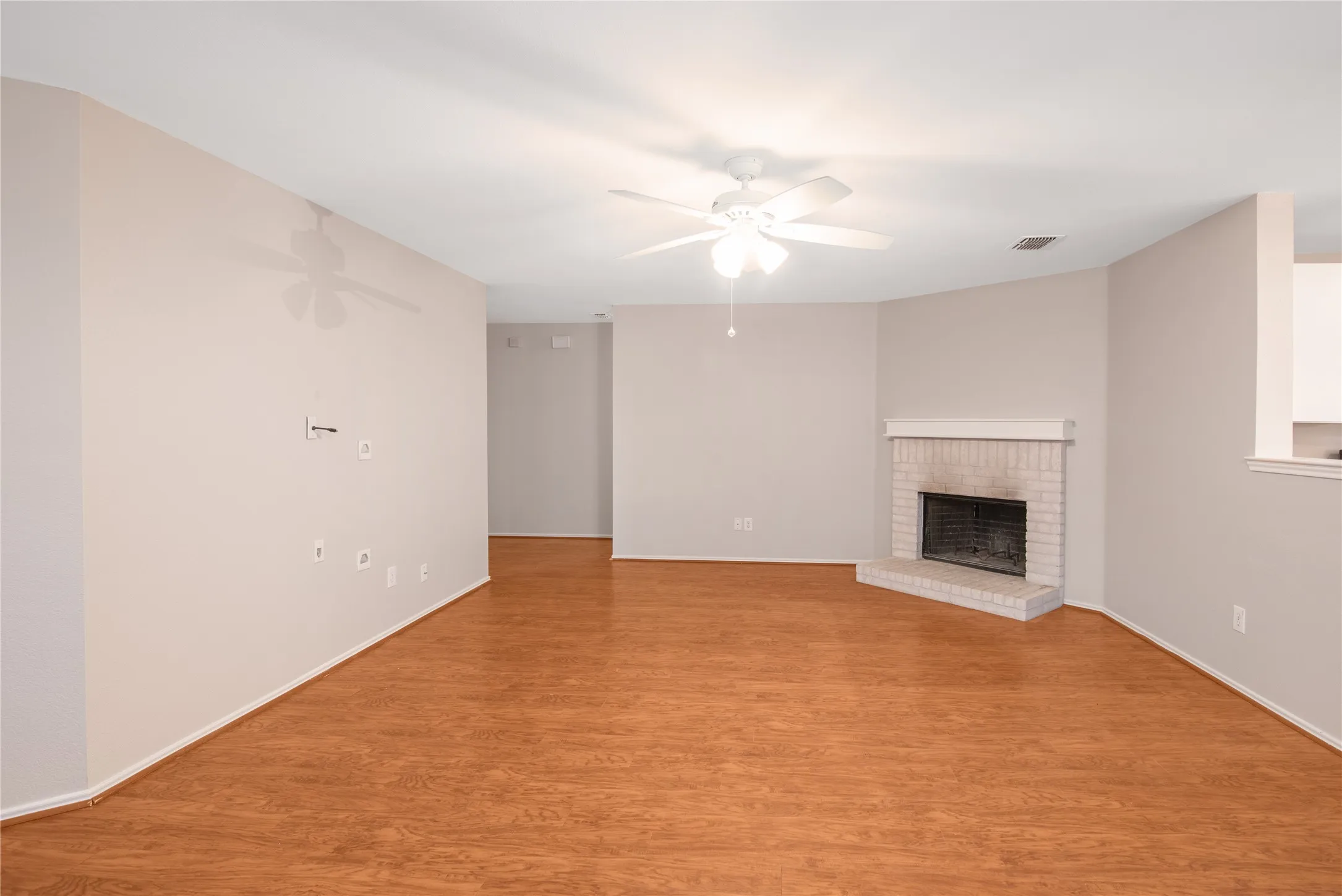 Spacious living area featuring wood-finish flooring, a white brick fireplace with mantel, and a ceiling fan