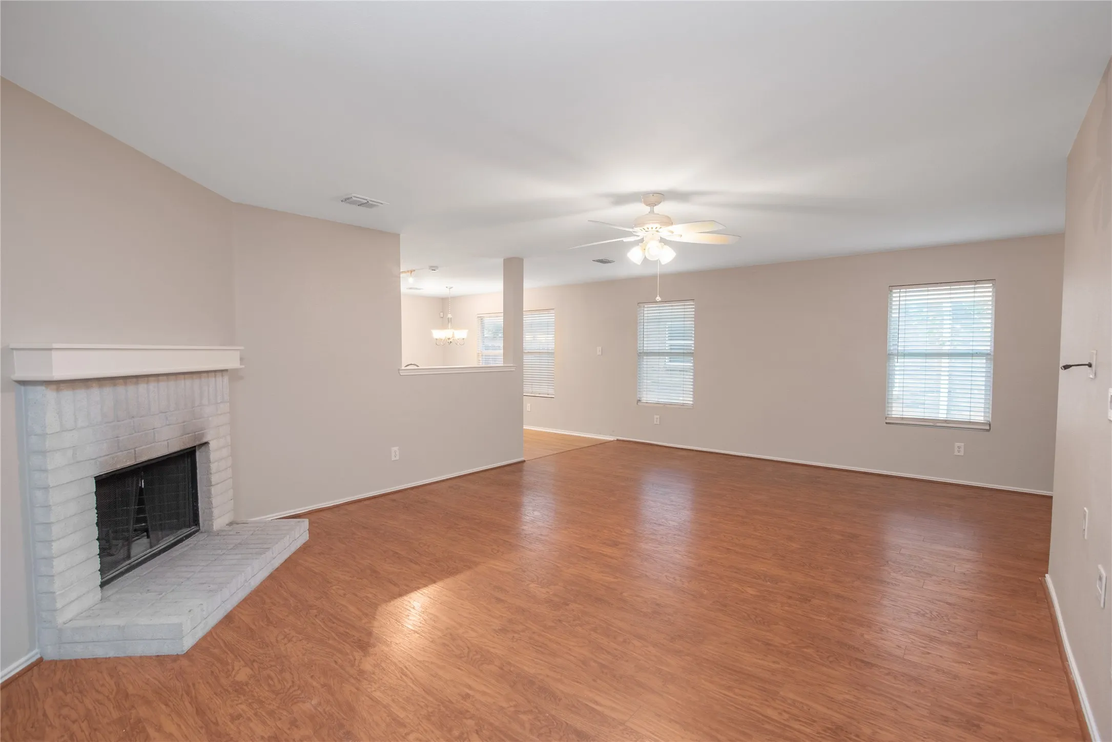 Expansive room featuring wood-finish flooring, a white brick fireplace with hearth, multiple windows, and a ceiling fan with integrated lighting