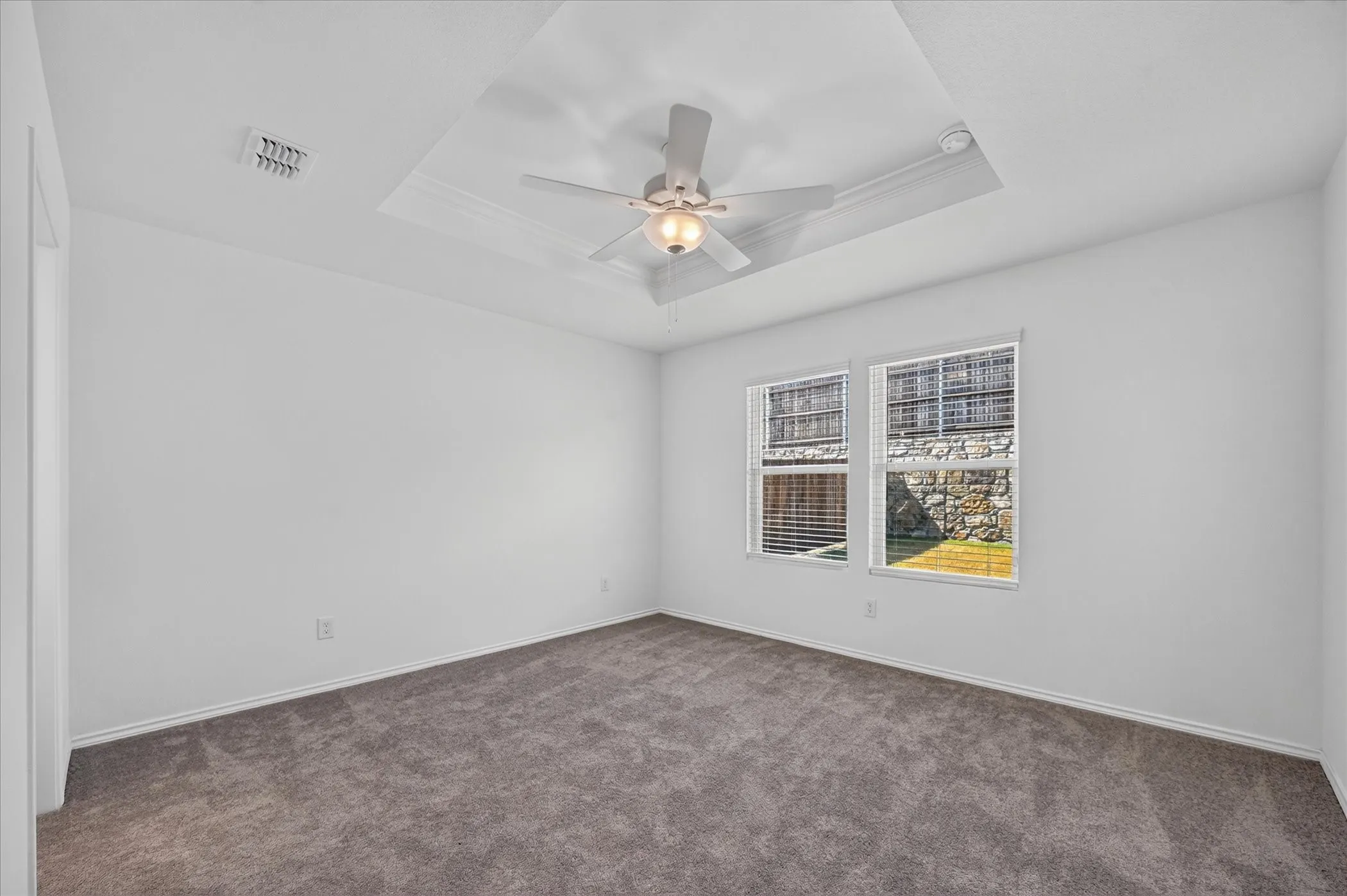 Tray ceiling with crown molding, ceiling fan with integrated lighting, dual pane windows with blinds, and neutral carpeting
