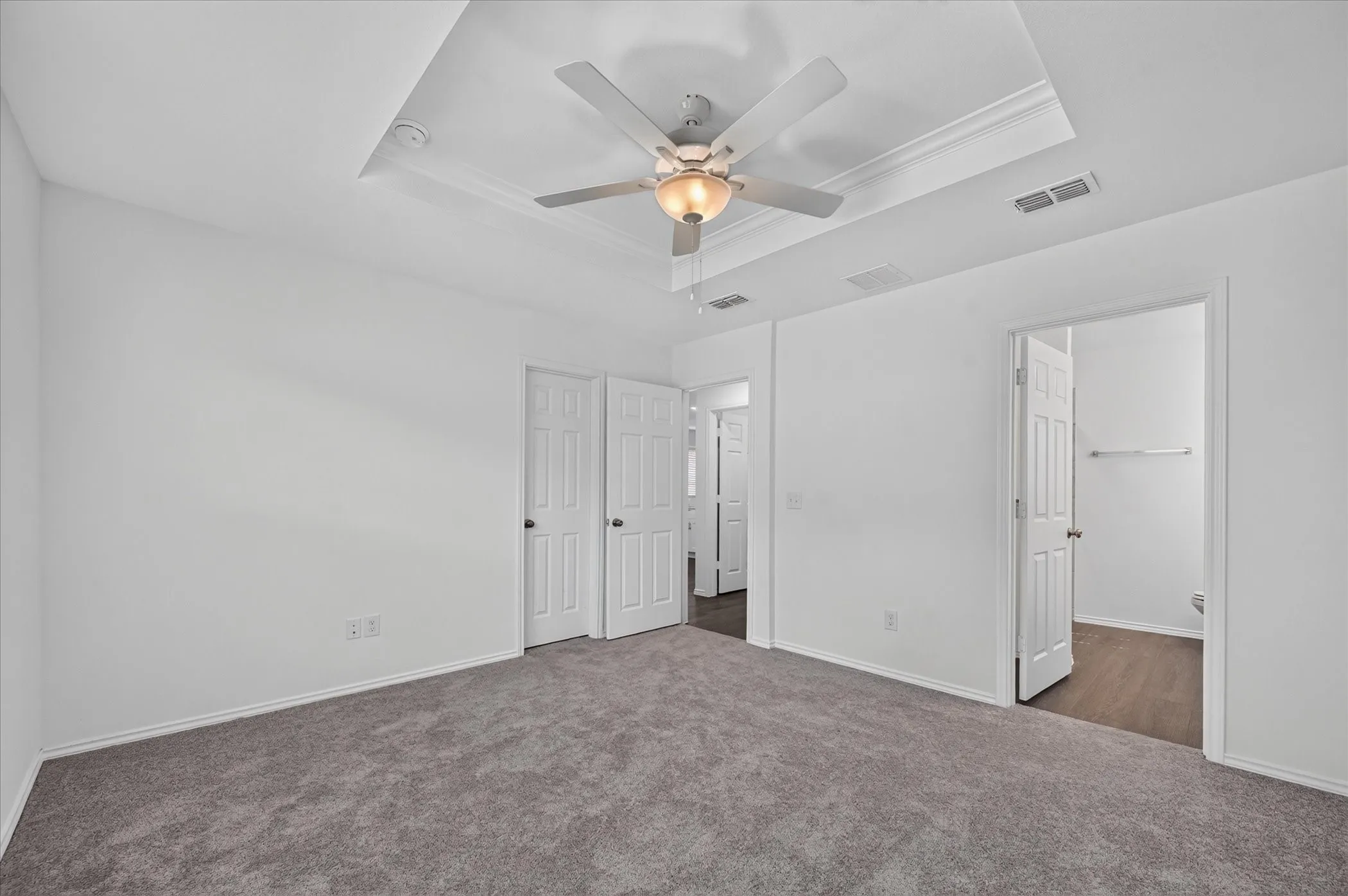 Tray ceiling with crown molding, ceiling fan with integrated lighting, gray carpet flooring, white paneled doors, and wood-finish flooring visible through doorway