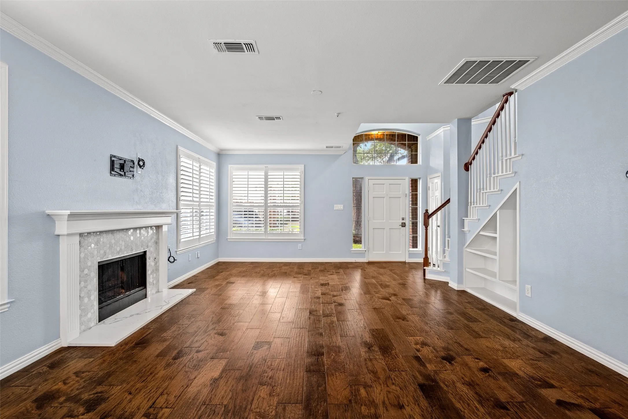 Wood-finish flooring, white fireplace mantel with tile surround, white plantation shutters, staircase with dark wood handrail, and built-in shelving under stairs