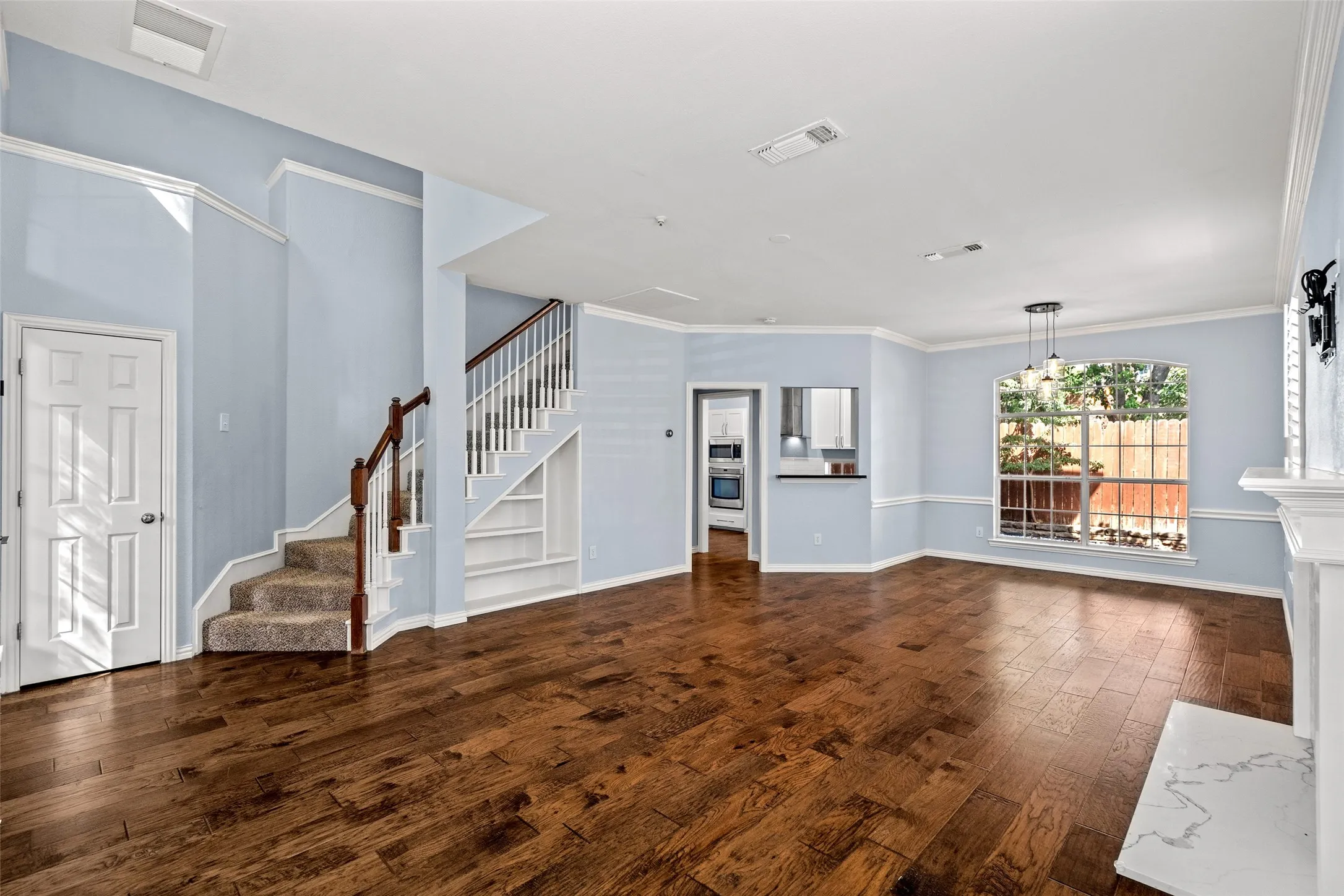 Hardwood-finish flooring throughout, white crown molding, built-in shelving under staircase, fireplace with a white mantel and light-toned hearth, and a window with garden scenery