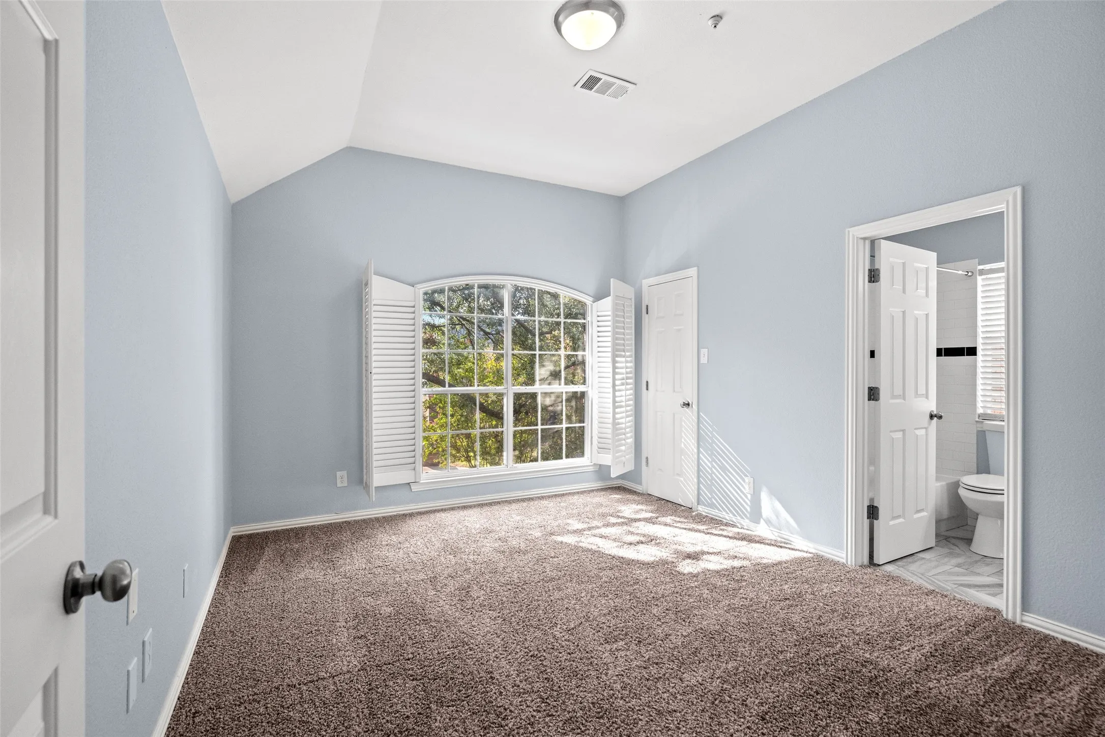 Vaulted ceiling, arched window with plantation shutters, plush carpeting, and an attached bathroom with tiled flooring
