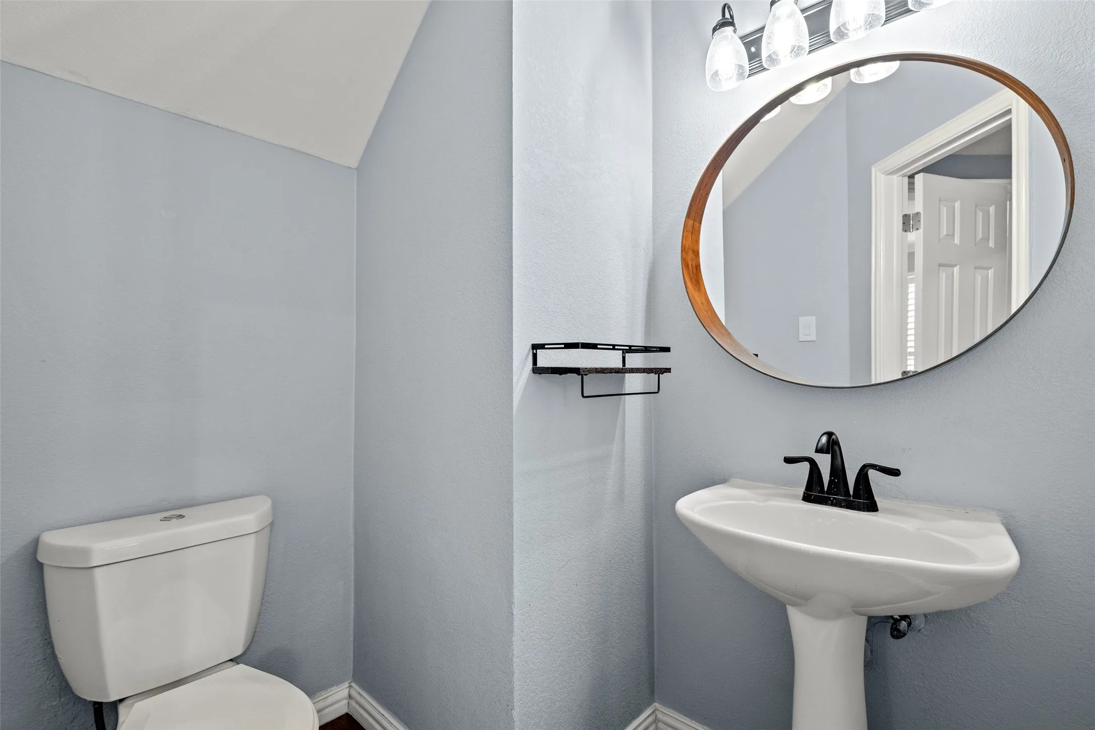 Pedestal sink, matte black faucet, wall-mounted oval mirror, three-light vanity fixture, and a toilet