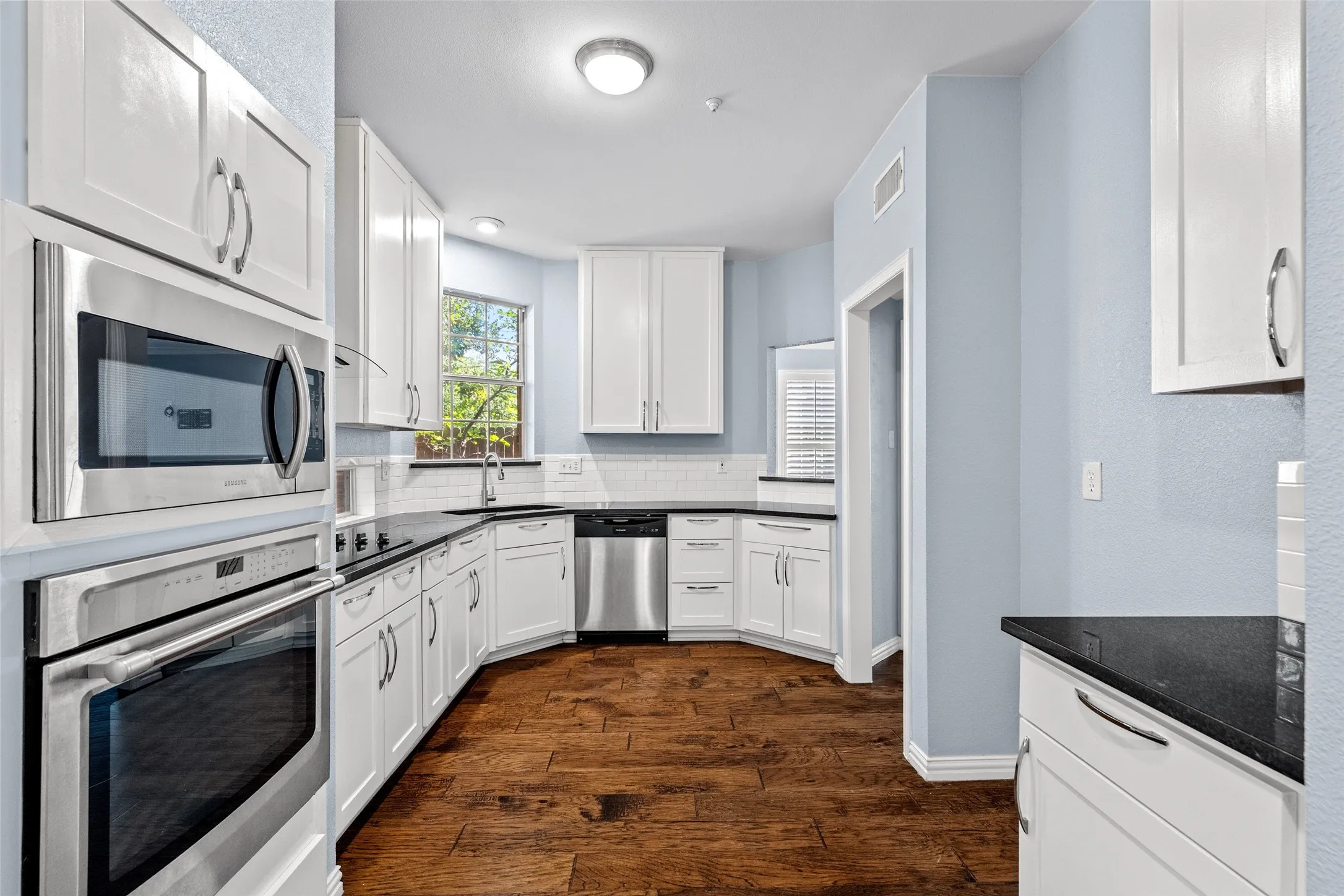Kitchen featuring white cabinetry, dark countertops, built-in stainless steel appliances, wood-finish flooring, and a white subway tile backsplash