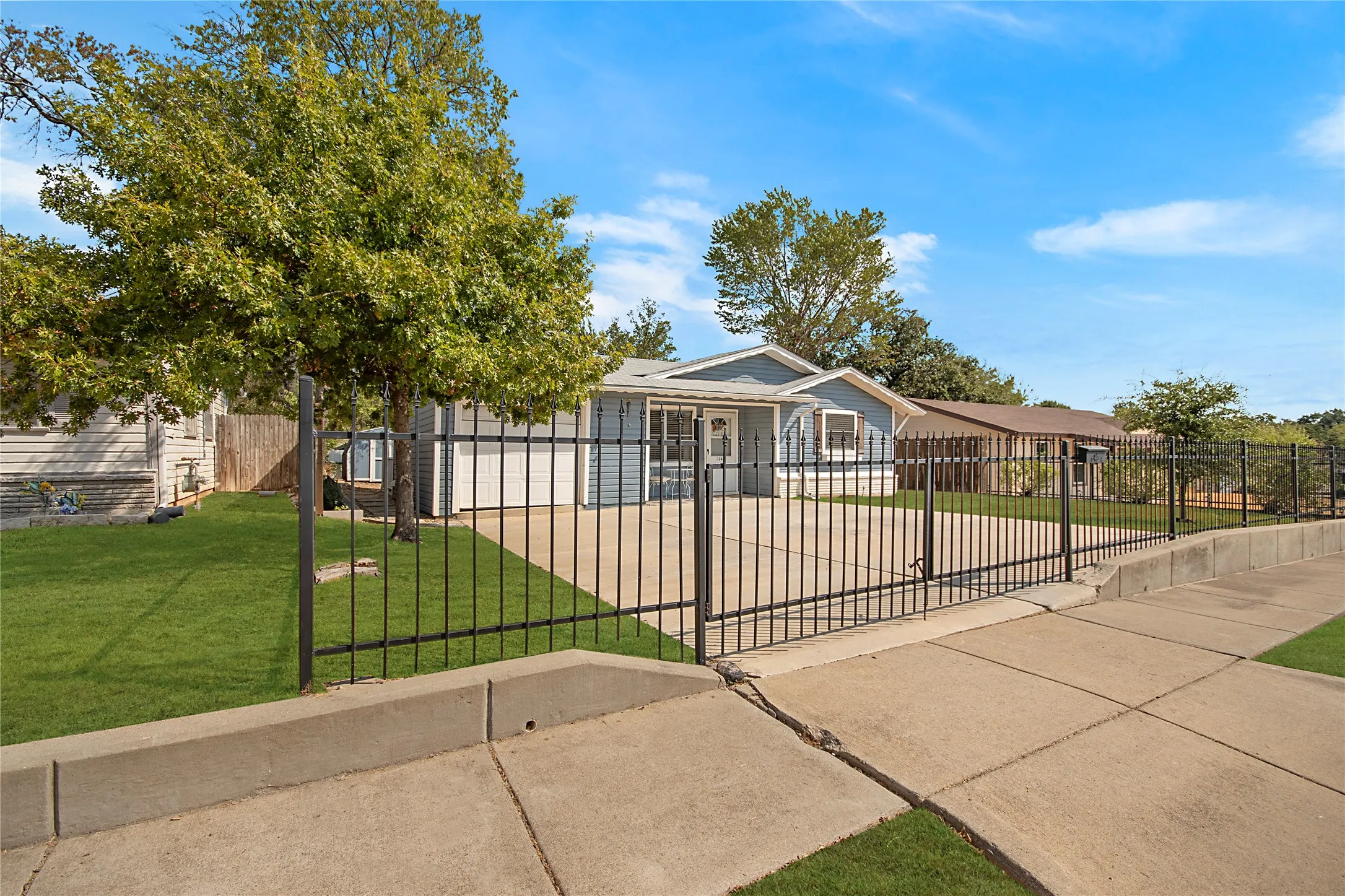 Fenced front yard with a concrete driveway, mature trees, and a light blue siding exterior