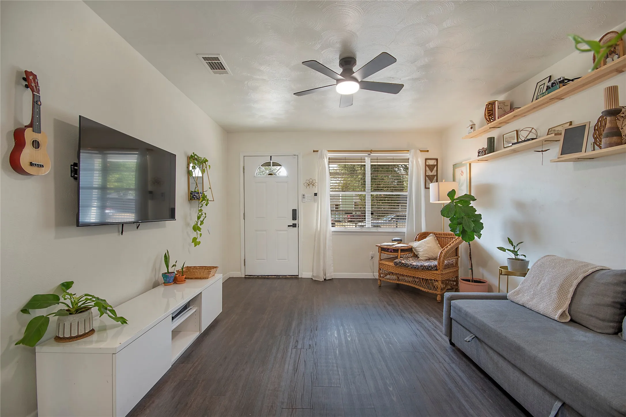 Wood-finish flooring throughout, ceiling fan with integrated lighting, white front door with decorative glass insert, and built-in floating shelves