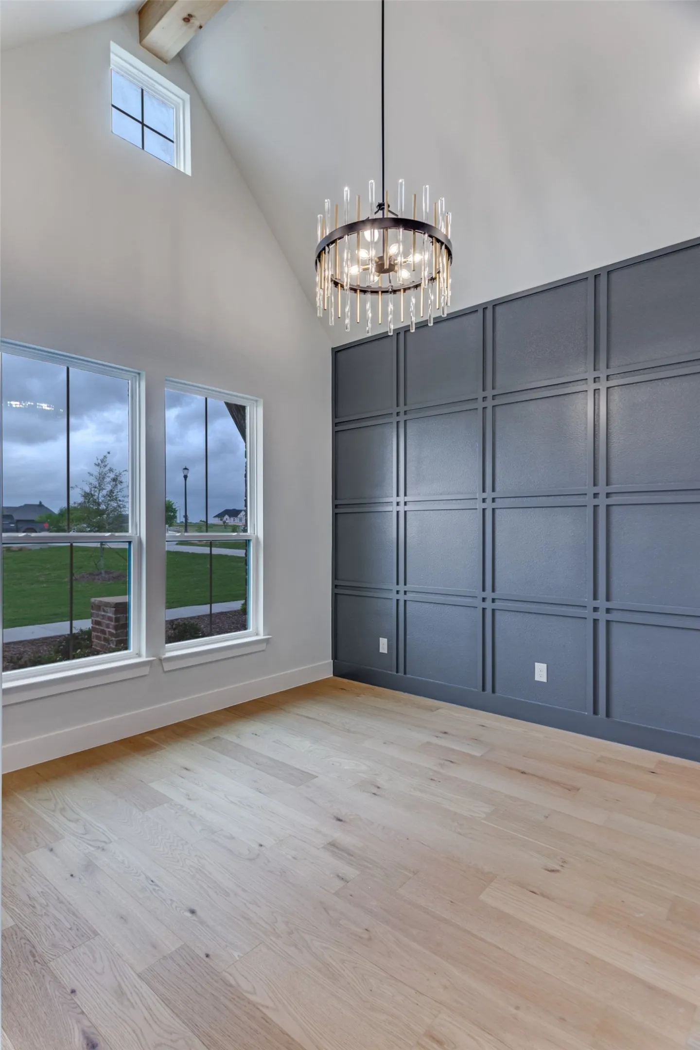 Vaulted ceiling with exposed wood beam, modern chandelier, two large windows, accent wall with panel molding, and wood-finish flooring