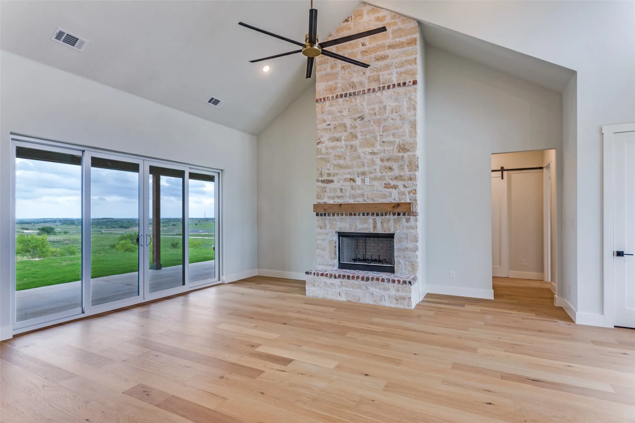 Vaulted ceiling with an exposed beam fan, stone fireplace with an integrated wood mantel, light wood-finish flooring, and multi-panel sliding glass doors