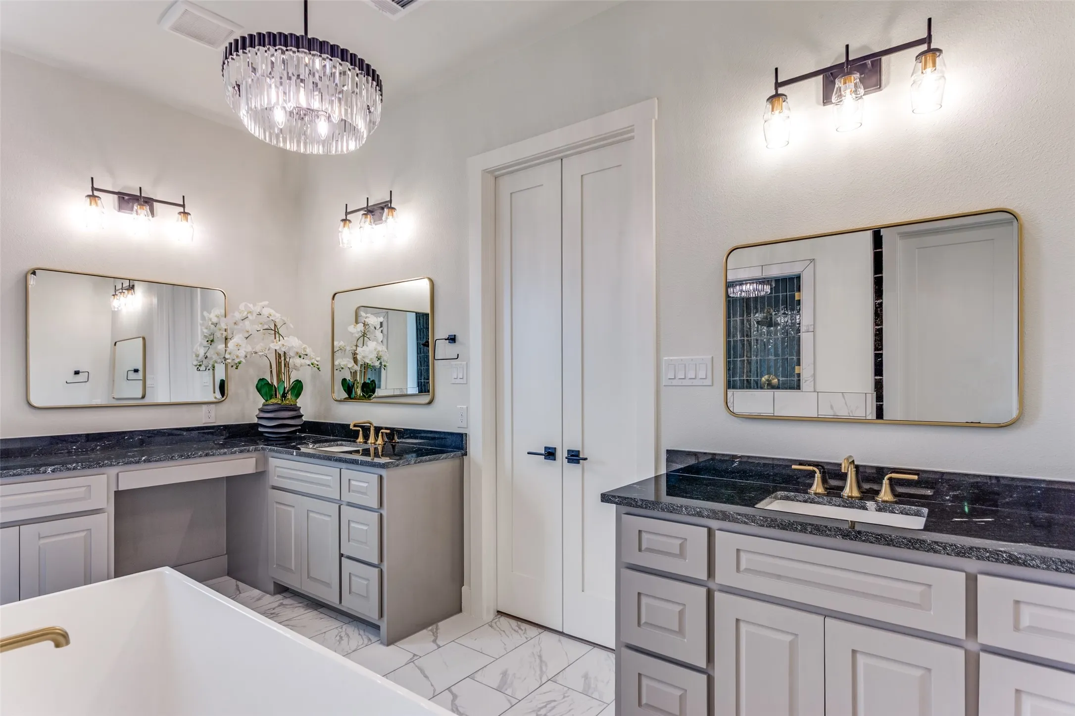 White tile flooring with grey veining, light grey cabinetry, dark countertops, a double vanity with undermount sinks, and gold-toned fixtures