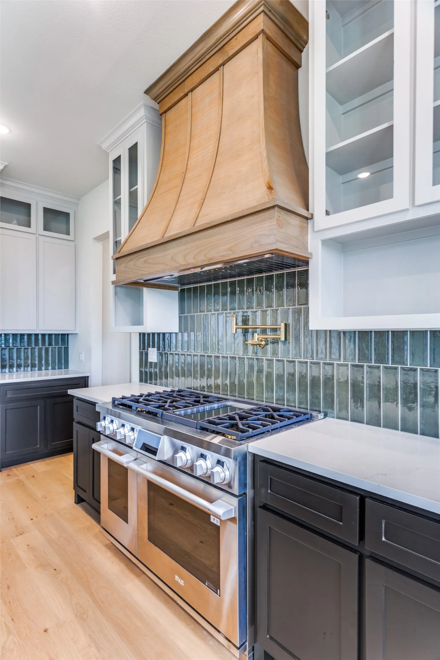 Stainless steel range with double ovens, wood-finish range hood, glass subway tile backsplash, light wood-finish flooring, and white countertops
