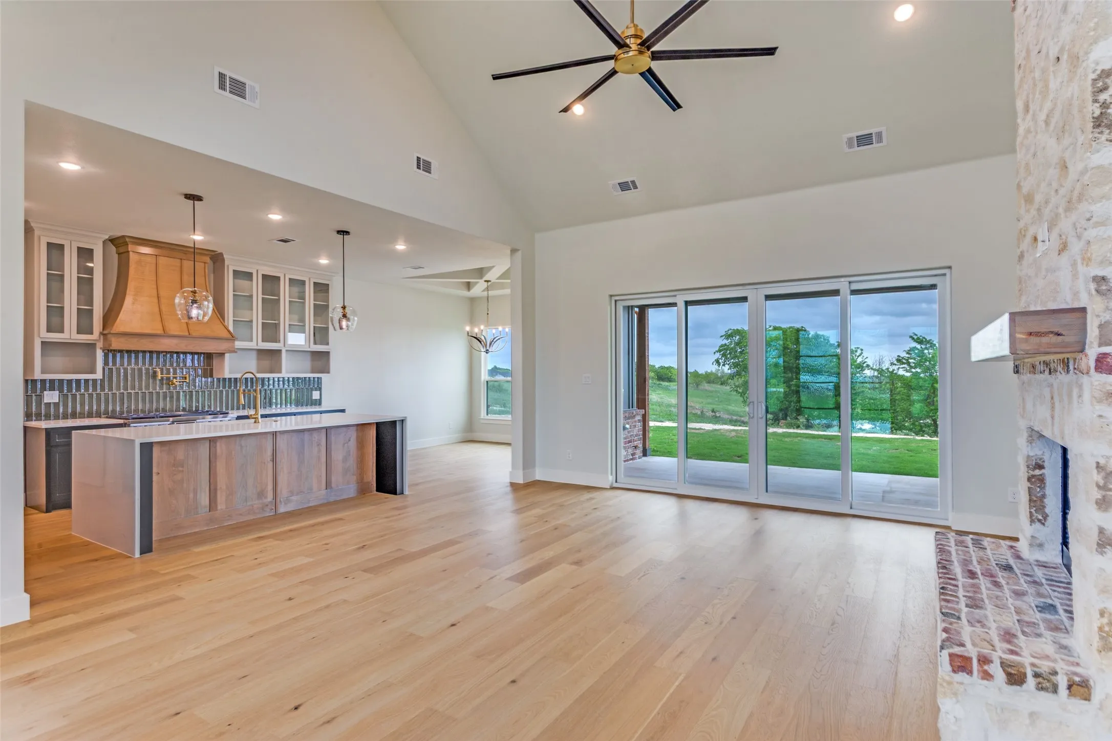 Vaulted ceiling with an exposed beam fan, light wood-finish flooring, stone fireplace with a wood mantle, custom kitchen island with a built-in sink, and full-wall sliding glass doors