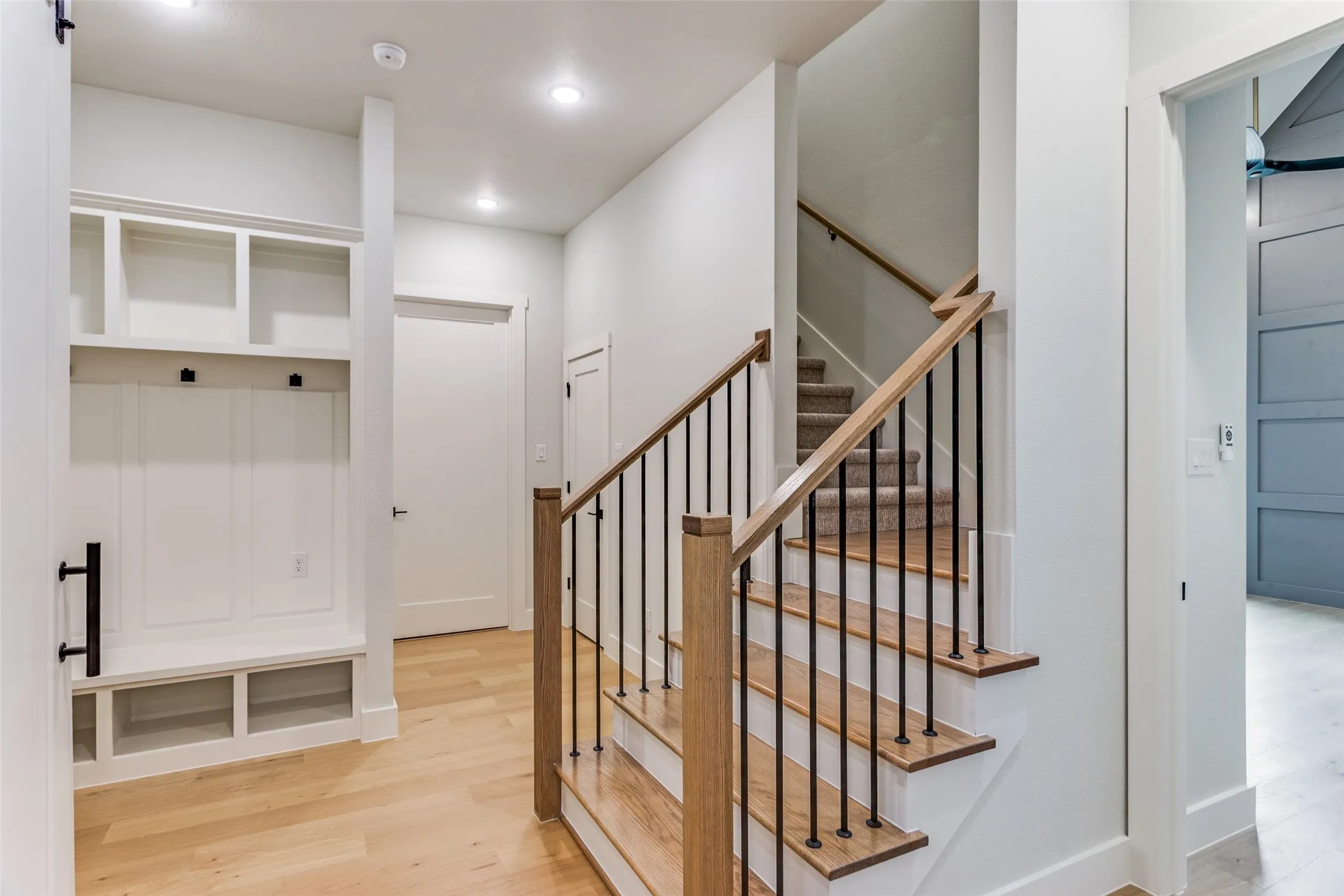 Built-in mudroom storage with hooks and cubbies, wood-finish flooring, staircase with wood banister and wrought iron balusters, recessed lighting, and a contemporary paneled door