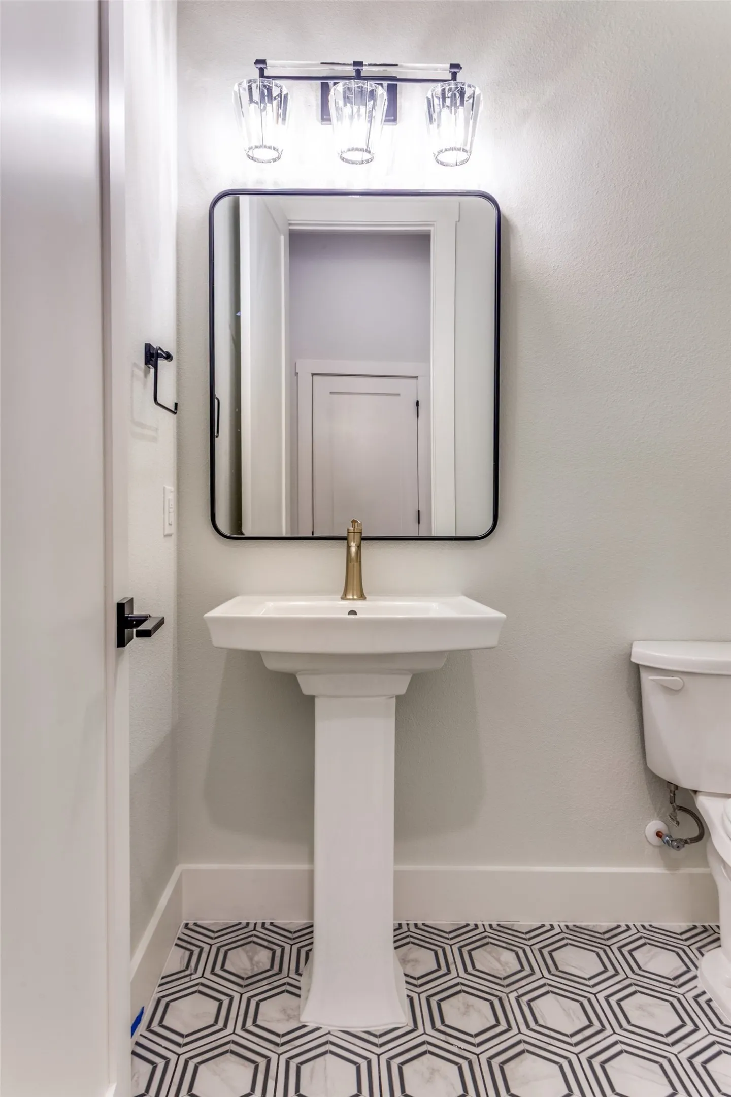 Pedestal sink with brass-finish faucet, framed mirror, three-light vanity fixture, and patterned hexagonal floor tile