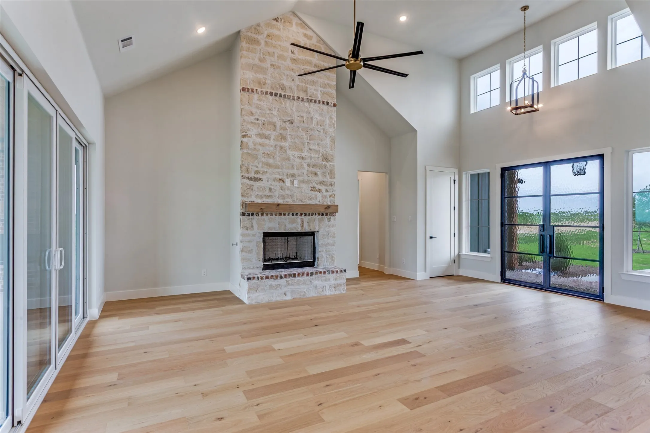 Vaulted ceilings, a floor-to-ceiling stone fireplace, light wood-finish flooring, black-framed glass doors, and a decorative chandelier