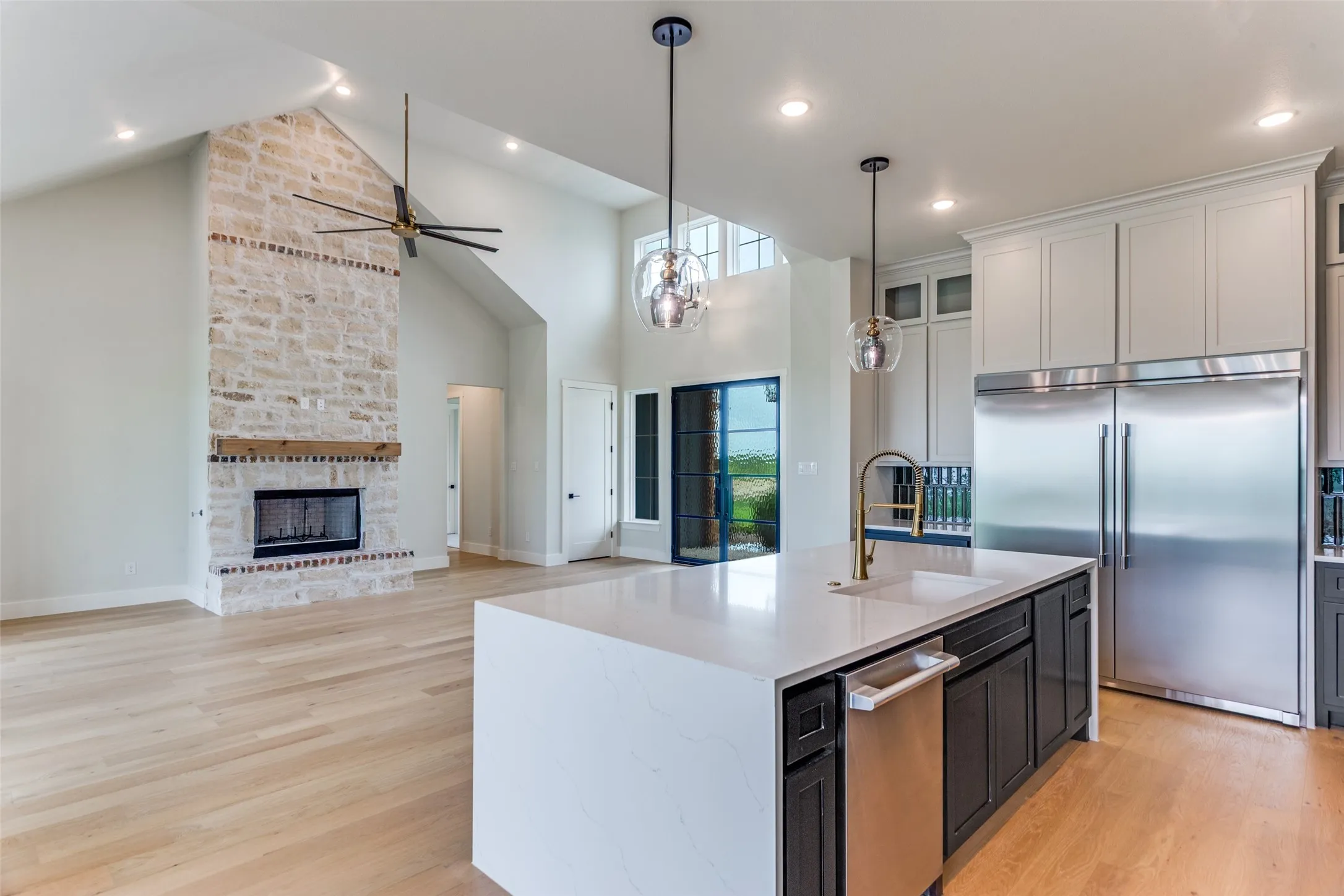 Vaulted ceilings, a stone fireplace with a wood-beam mantel, wood-finish flooring, a kitchen island with sink, and stainless steel appliances