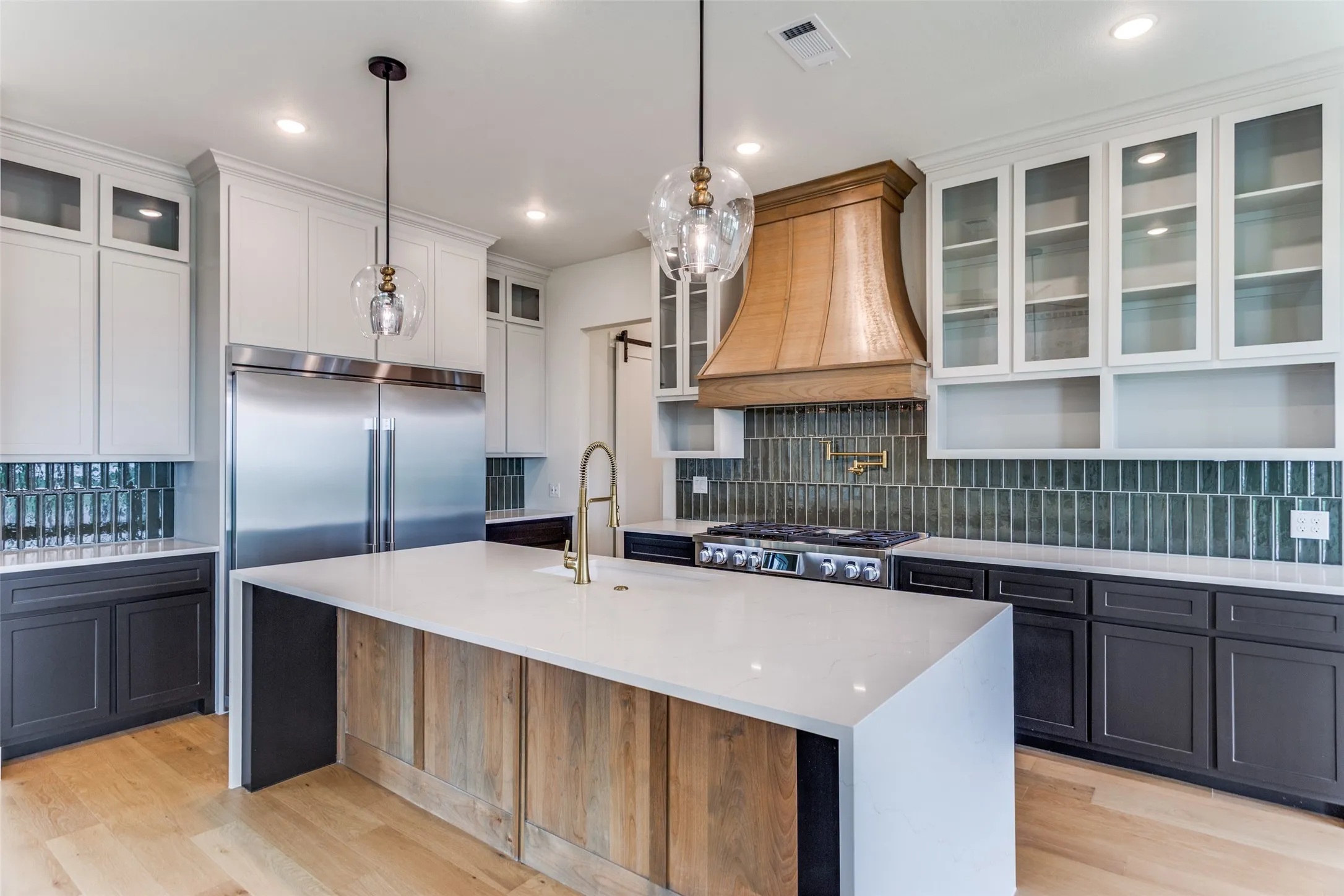 Kitchen featuring a large central island with a light-toned countertop, stainless steel appliances, wood-finish flooring, a custom wood range hood, and a full-height tile backsplash