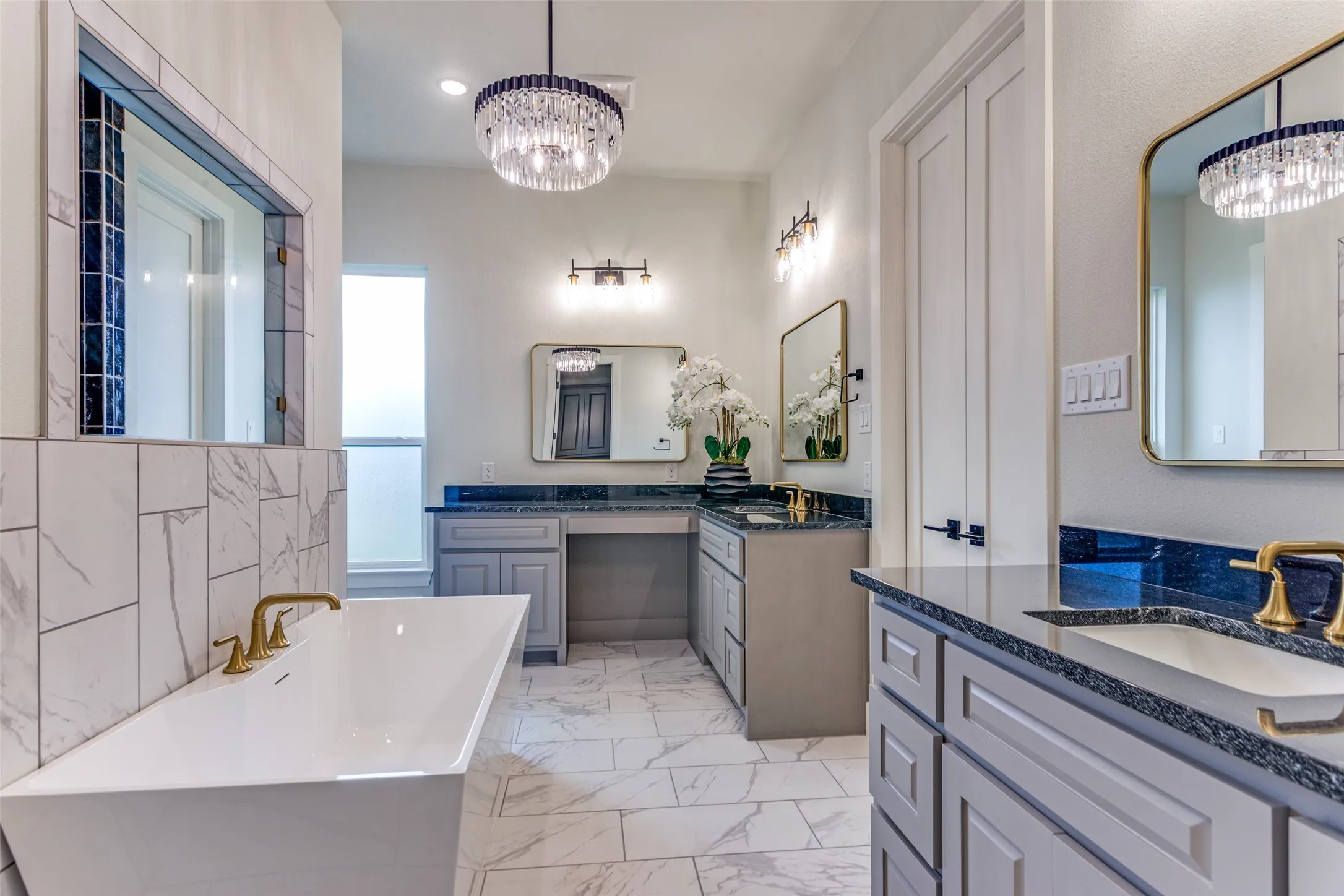 Freestanding soaking tub with brass fixtures, dual vanity with undermount sinks, stone countertops, and decorative lighting fixtures