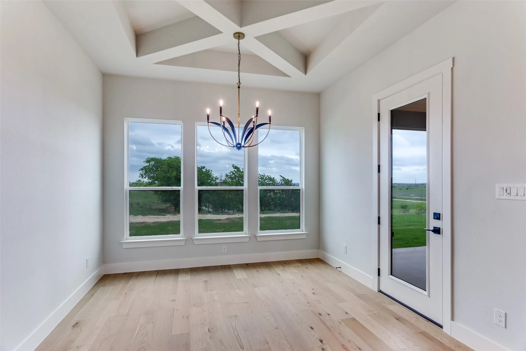 Wood-finish flooring, vaulted ceiling with exposed beams, three large windows, glass-paneled exterior door, and a decorative chandelier