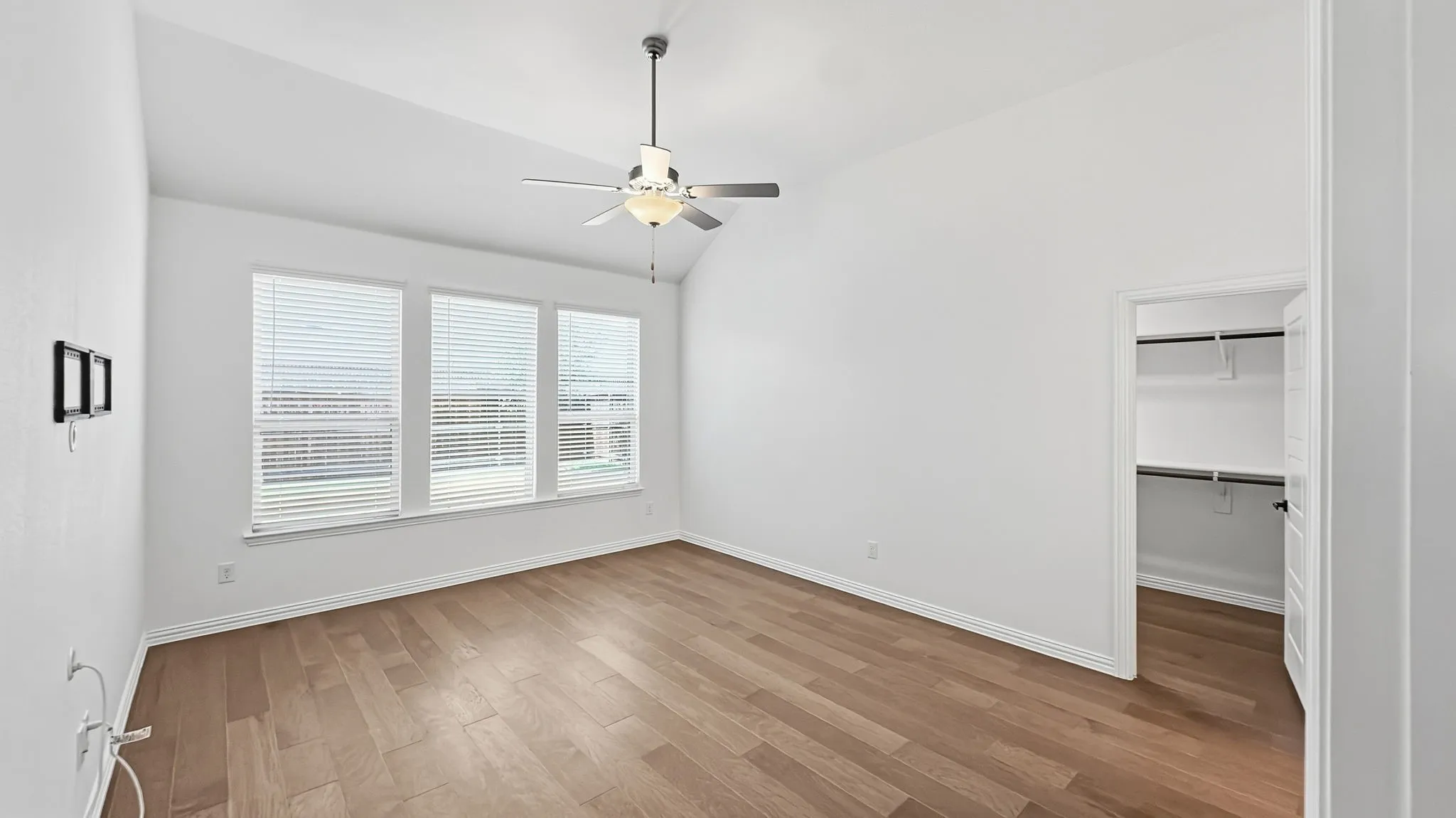 Vaulted ceiling with an attached ceiling fan, large windows with blinds, wood-finish flooring, and built-in closet shelving