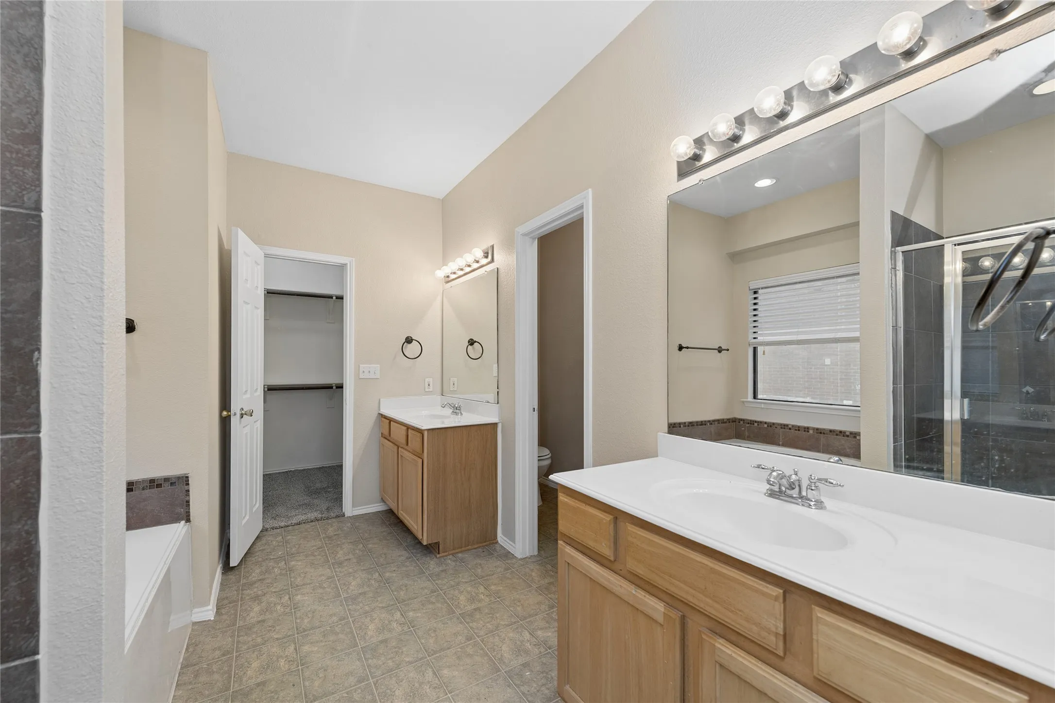 Dual vanity sinks with wood-finish cabinetry, tile flooring, glass-enclosed shower, and built-in bathtub