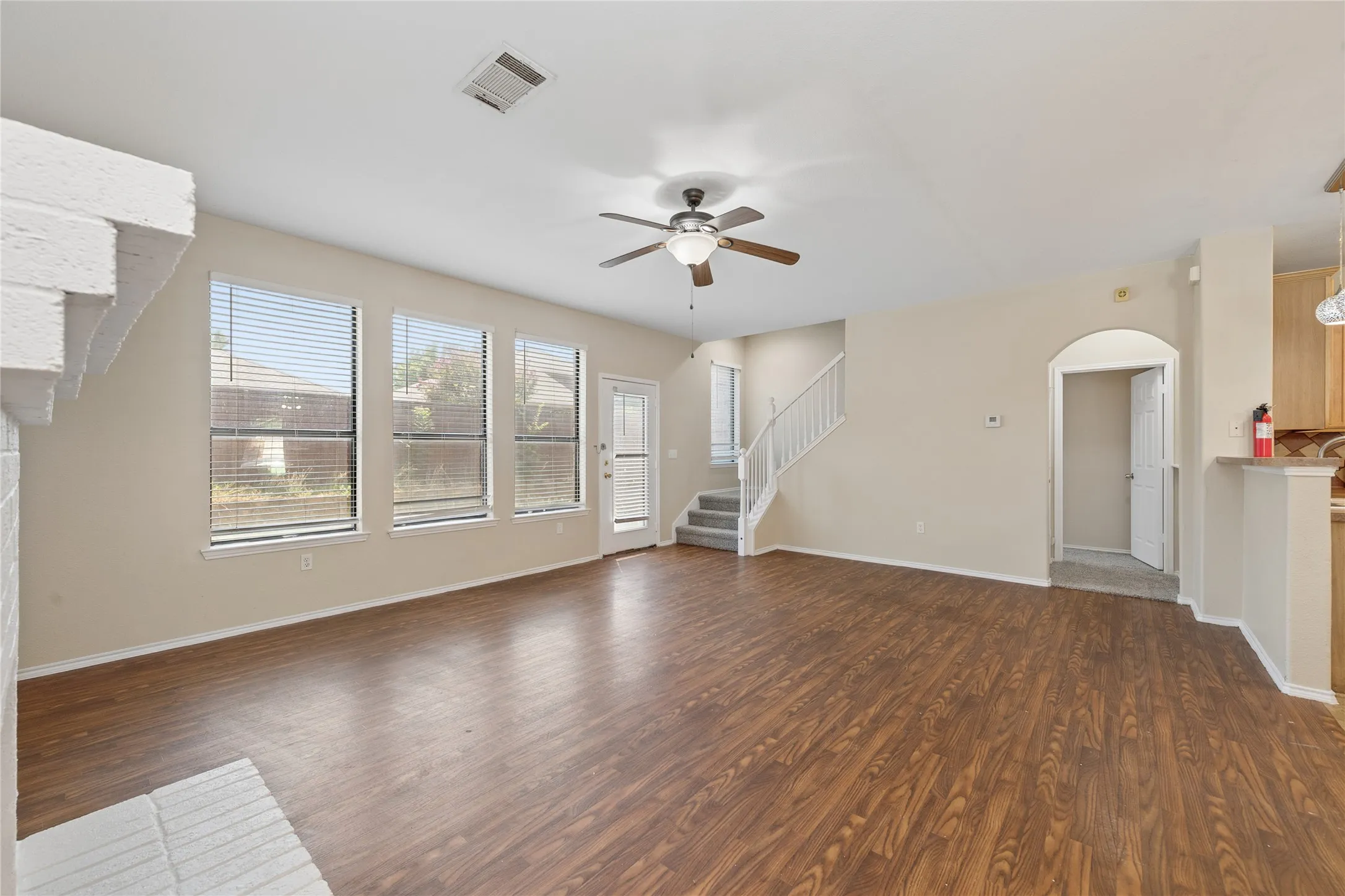 Spacious living area featuring wood-finish flooring, a ceiling fan, three windows with blinds, a white brick fireplace surround, and a staircase with white railings