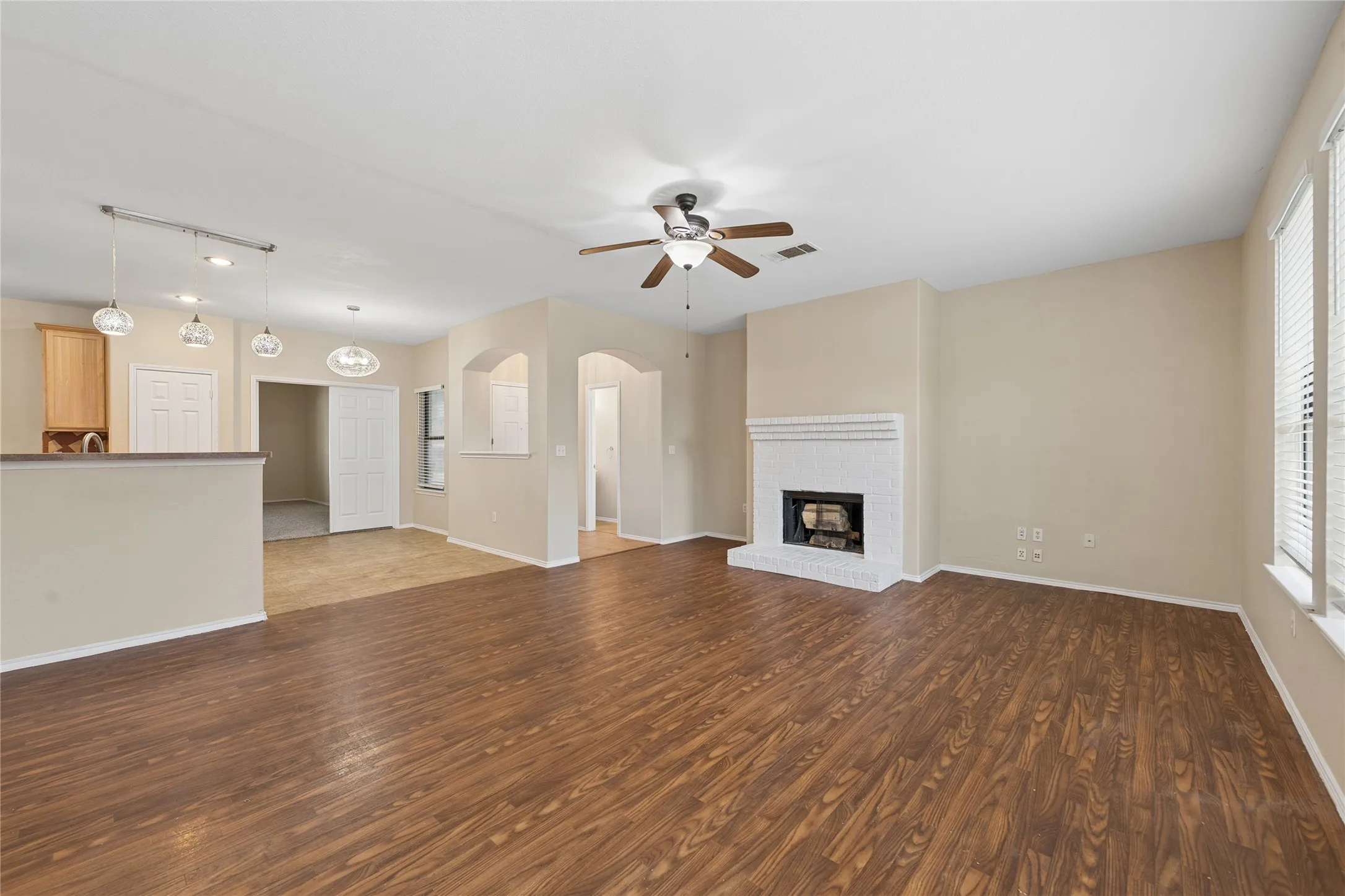 Wood-finish flooring, white brick fireplace, ceiling fan, multiple light fixtures, and archways