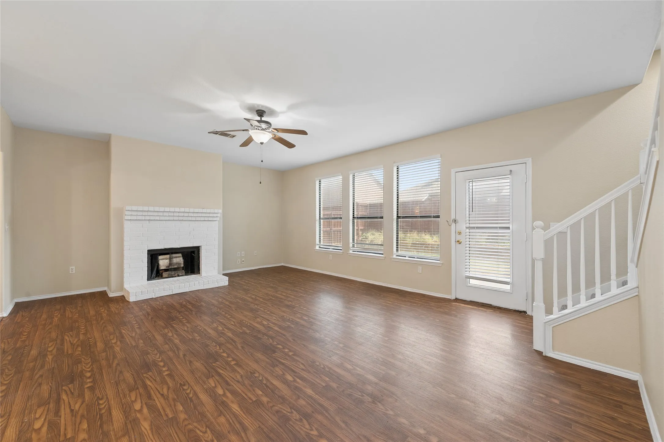 Wood-finish flooring, white brick fireplace, ceiling fan with light fixture, multiple windows, and a glass-paneled door