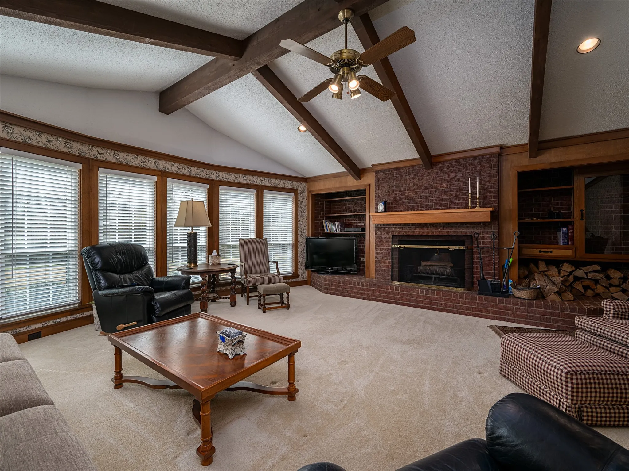 Vaulted ceiling with exposed wood beams, brick fireplace with wood mantel, built-in shelving, and multiple windows