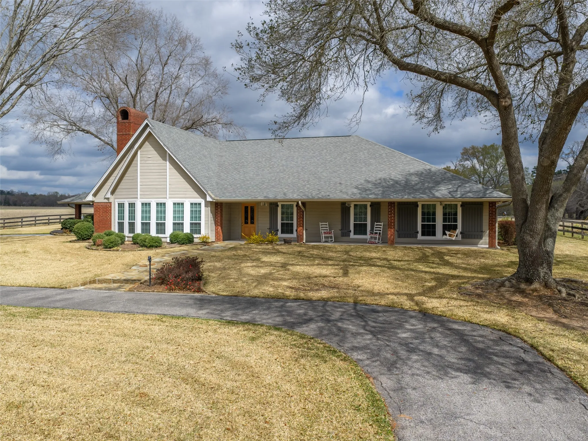 Gable roofline with a prominent brick chimney, large front windows, covered porch with columns, established trees, and a paved driveway