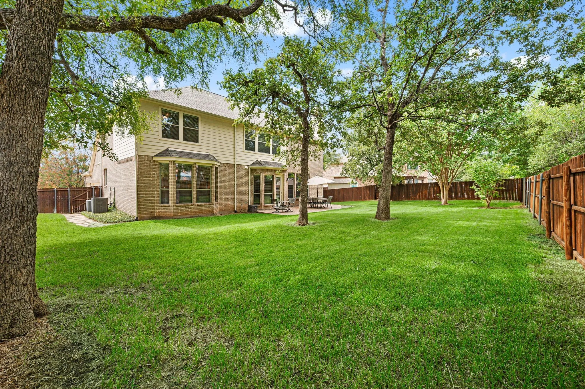 Expansive green lawn with mature trees, a two-story residence featuring a brick and siding exterior, multiple bay windows, and a fenced perimeter
