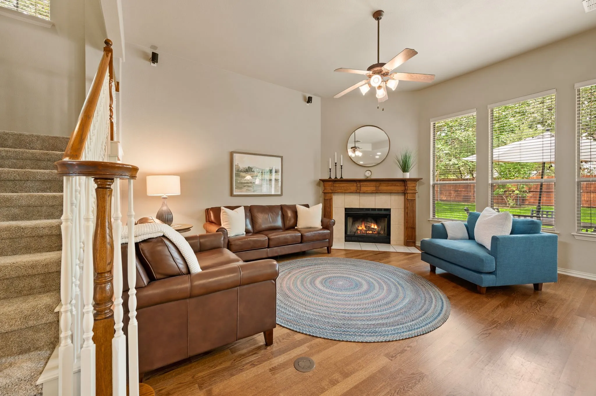 Hardwood-style flooring, fireplace with wood mantel, ceiling fan with integrated lighting, large windows, and carpeted staircase