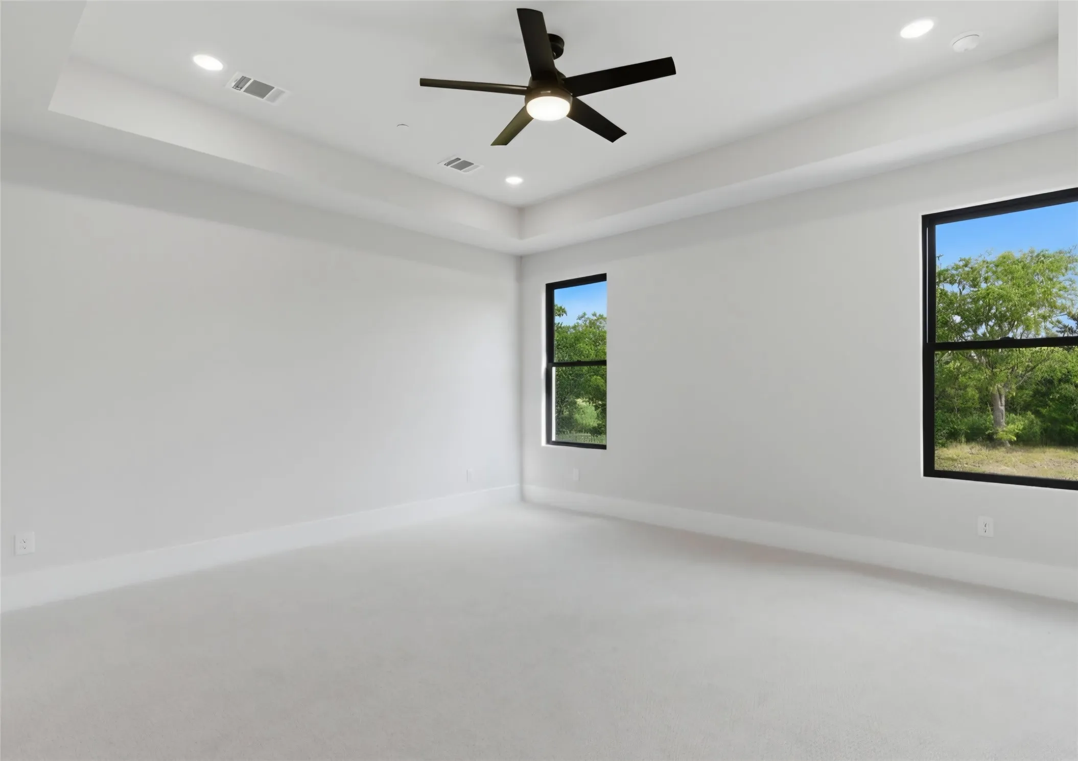 Recessed lighting, ceiling fan, tray ceiling, two windows with black frames, and light-toned carpet