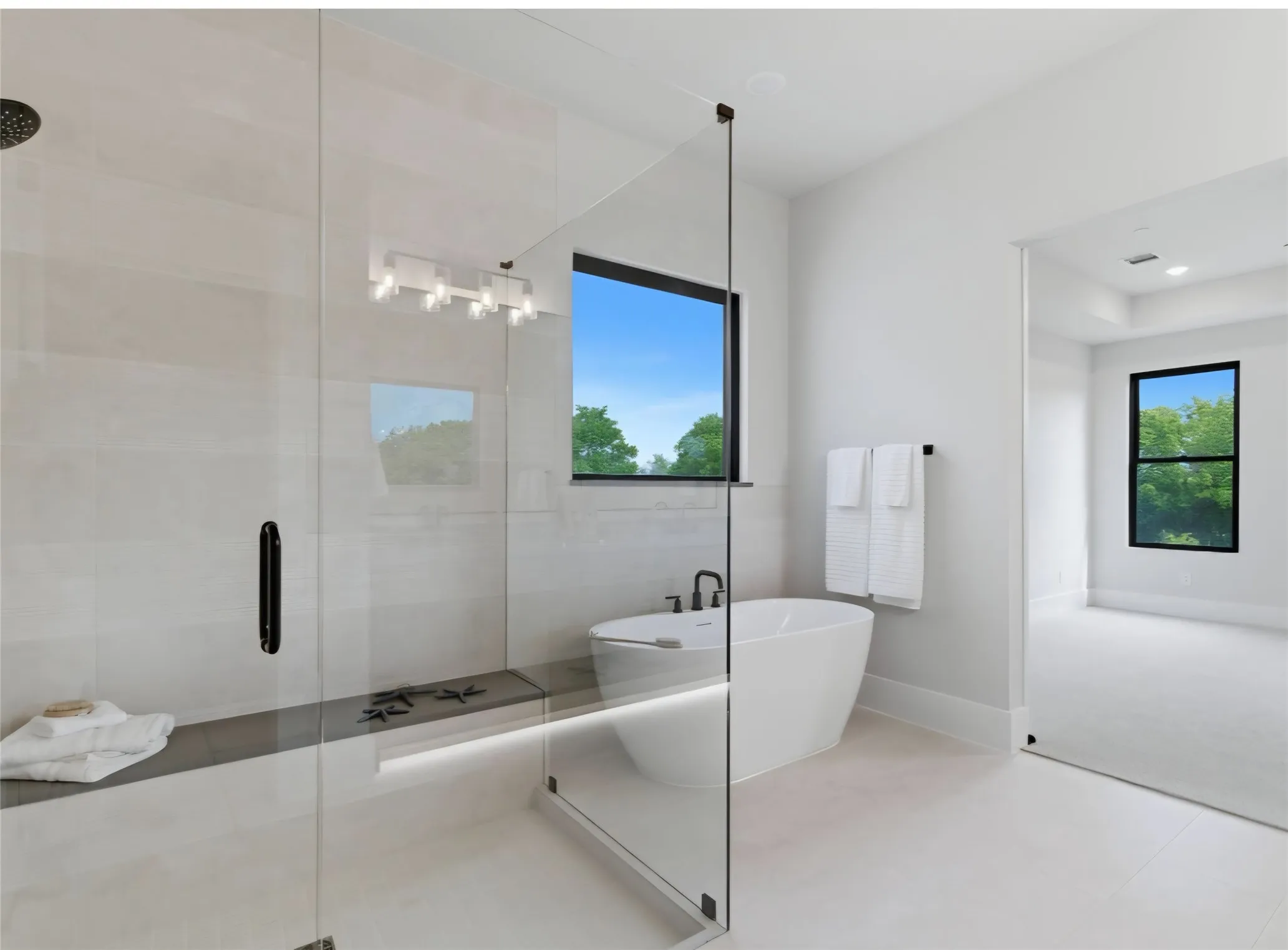 Freestanding soaking tub, glass-enclosed shower with tiled walls, built-in shower bench, and contemporary vanity lighting