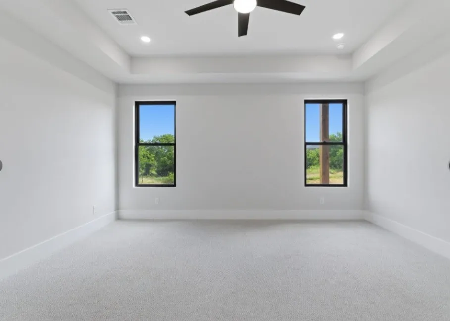 Recessed lighting and a ceiling fan illuminate this carpeted room, featuring two tall windows with dark frames and a tray ceiling