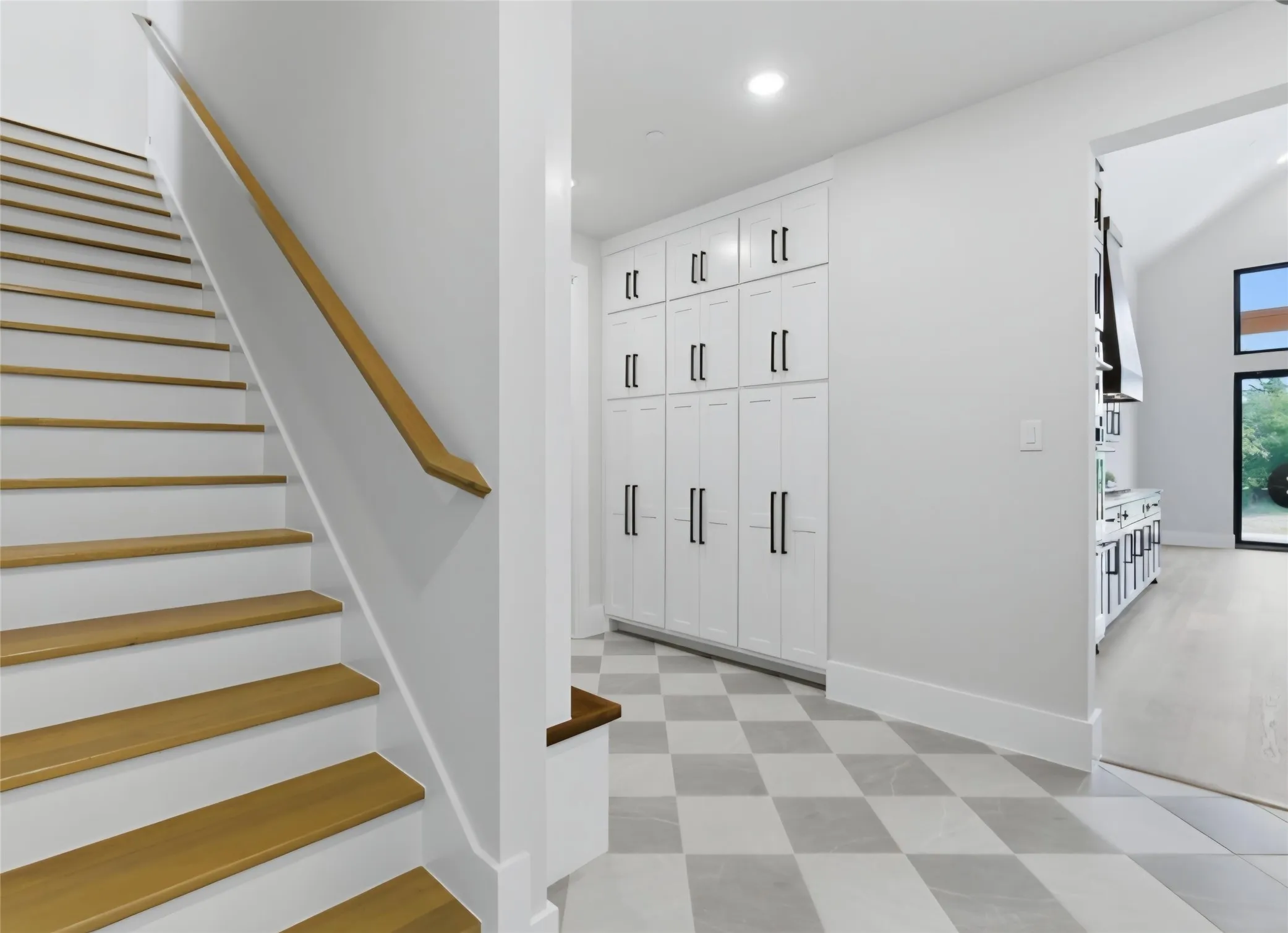Hardwood staircase with a wood-finish handrail, checkered tile flooring, built-in white cabinetry with matte black hardware, and recessed lighting