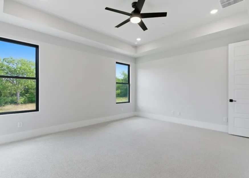Recessed lighting, ceiling fan, tray ceiling, two large windows with black trim, and light-colored carpeting