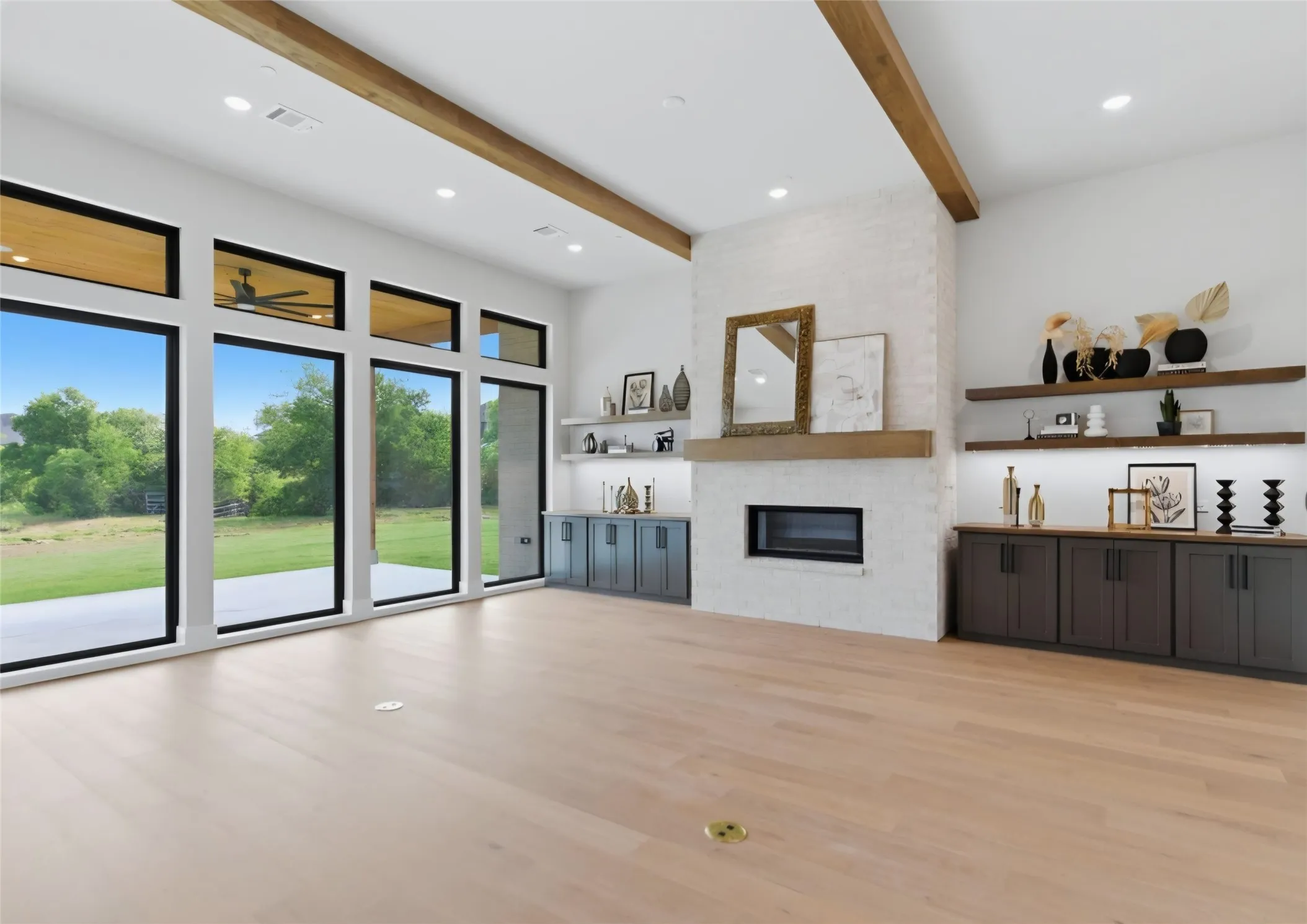 Open-concept living space with full-height windows, wood-finish flooring, a white brick fireplace, exposed ceiling beams, and built-in cabinetry with floating shelves
