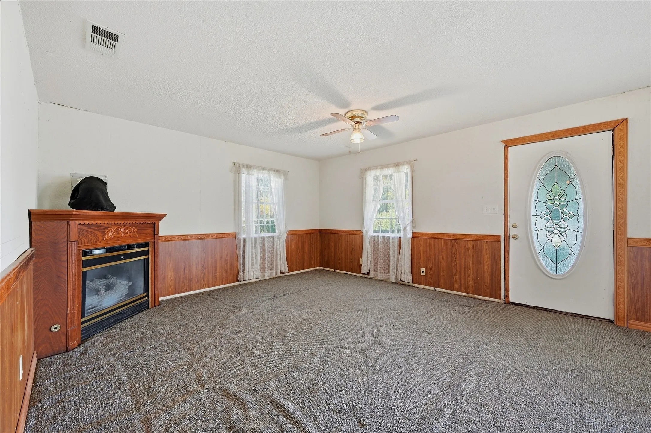 Carpeted living area featuring a decorative electric fireplace, wood-paneled wainscoting, two windows, and a ceiling fan