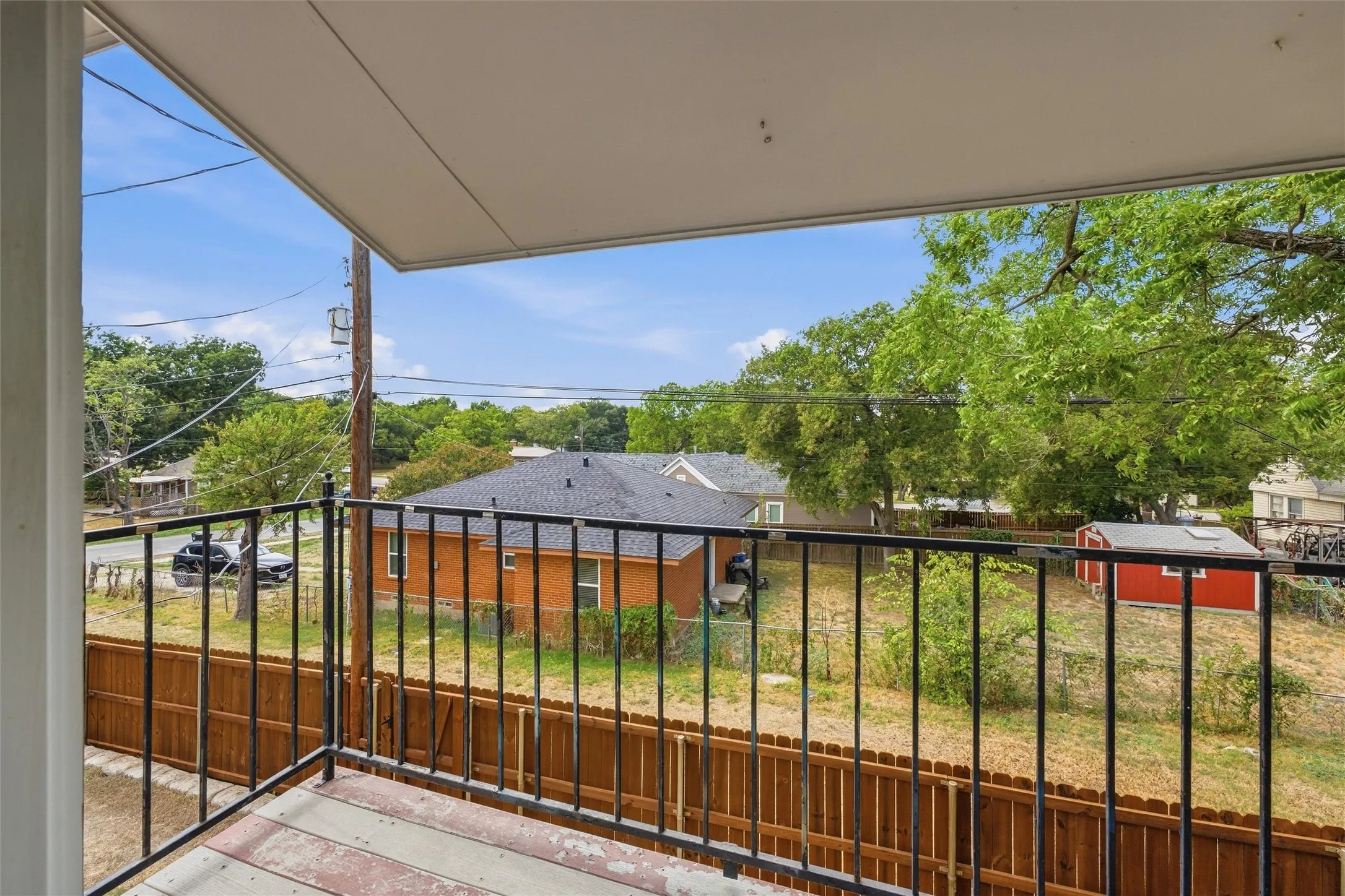 Balcony with black metal railing overlooking a fenced yard with mature trees