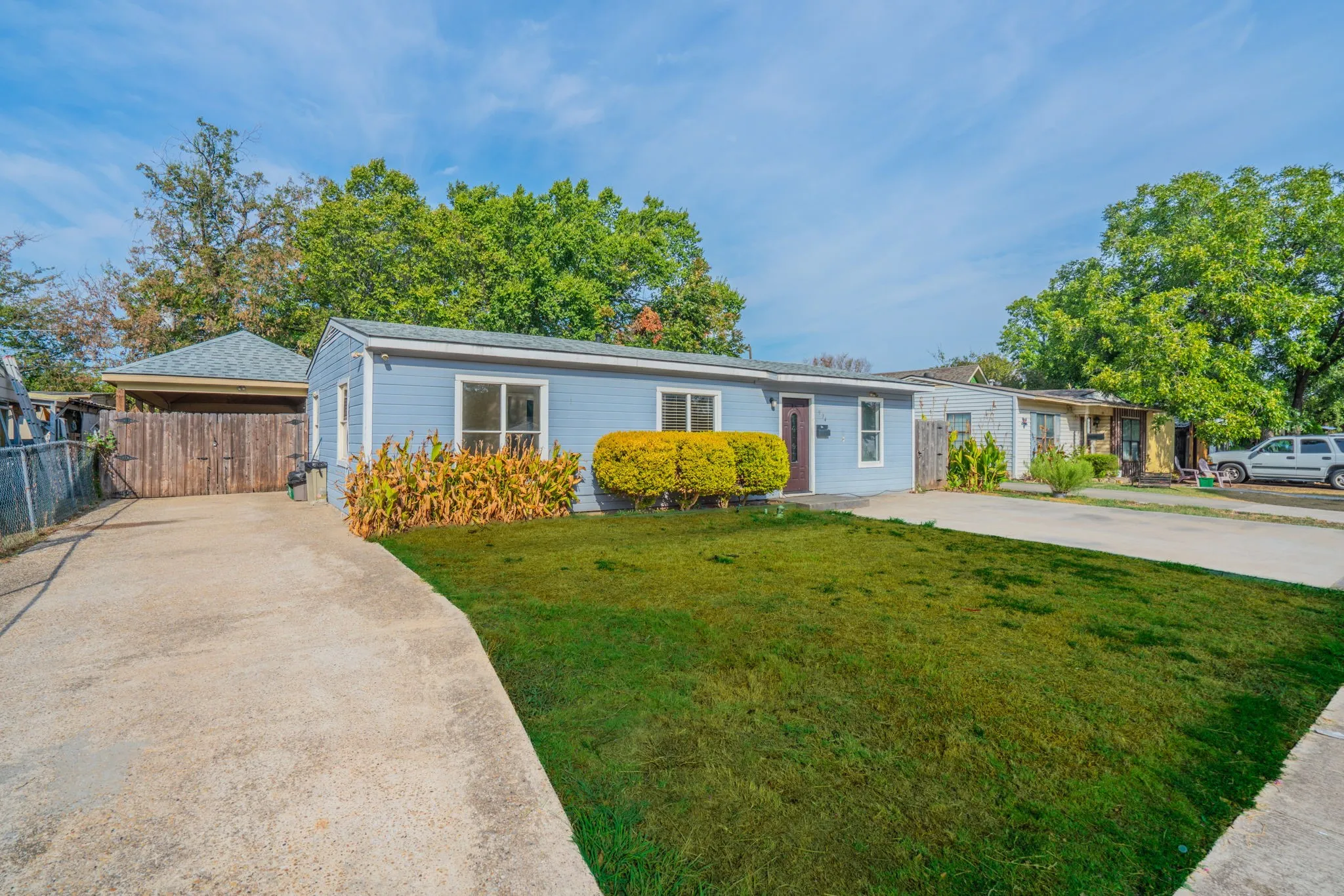 Light blue exterior siding, white window trim, front lawn, concrete driveway, and covered carport with wood fencing
