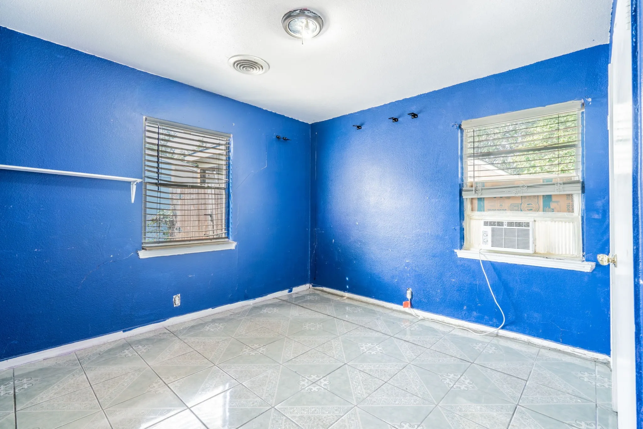 Tile flooring, two windows with blinds, wall-mounted air conditioning unit, ceiling-mounted light fixtures